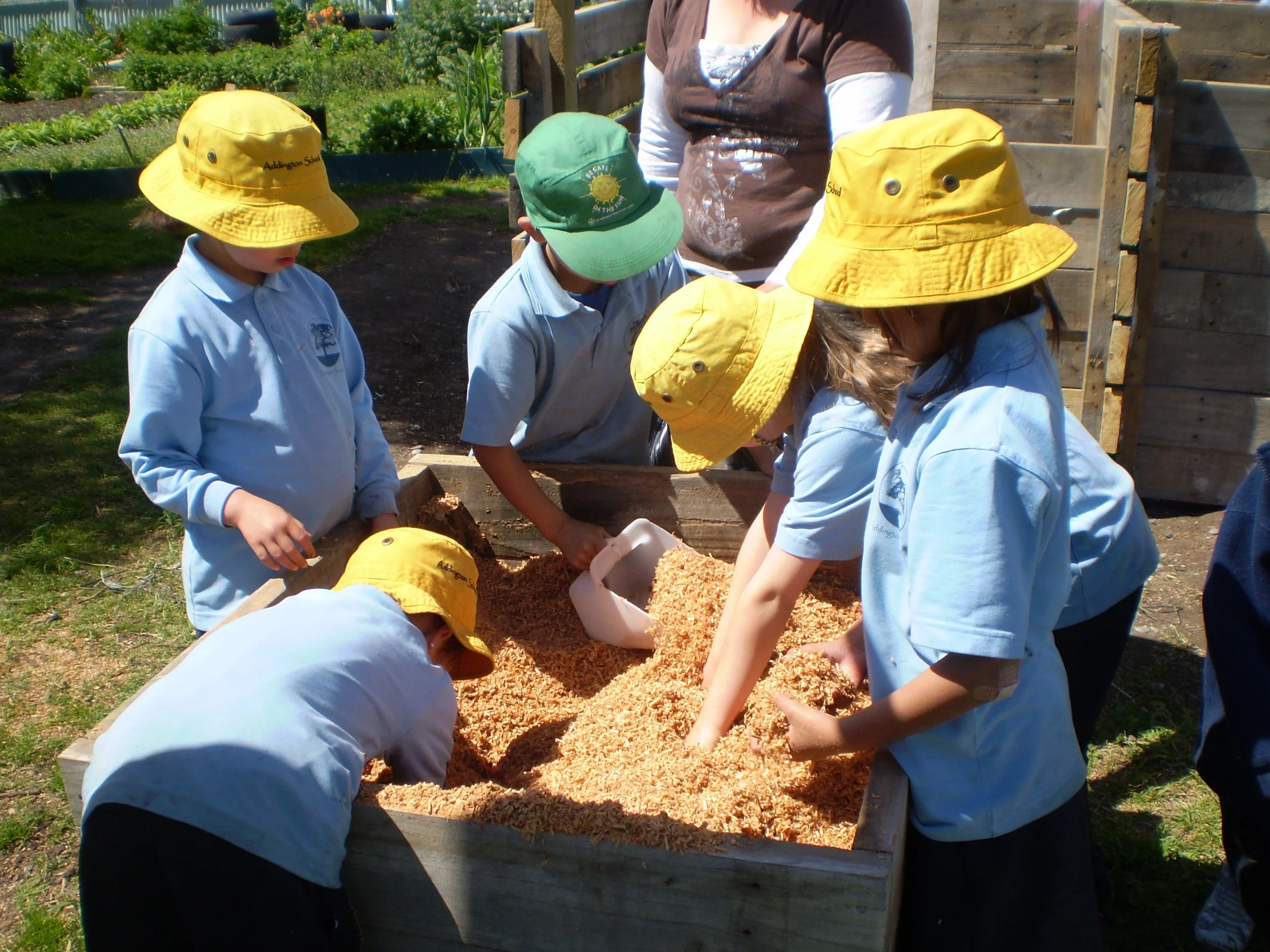 Group of children wearing yellow and green hats and blue uniforms playing in a sandbox at a garden.