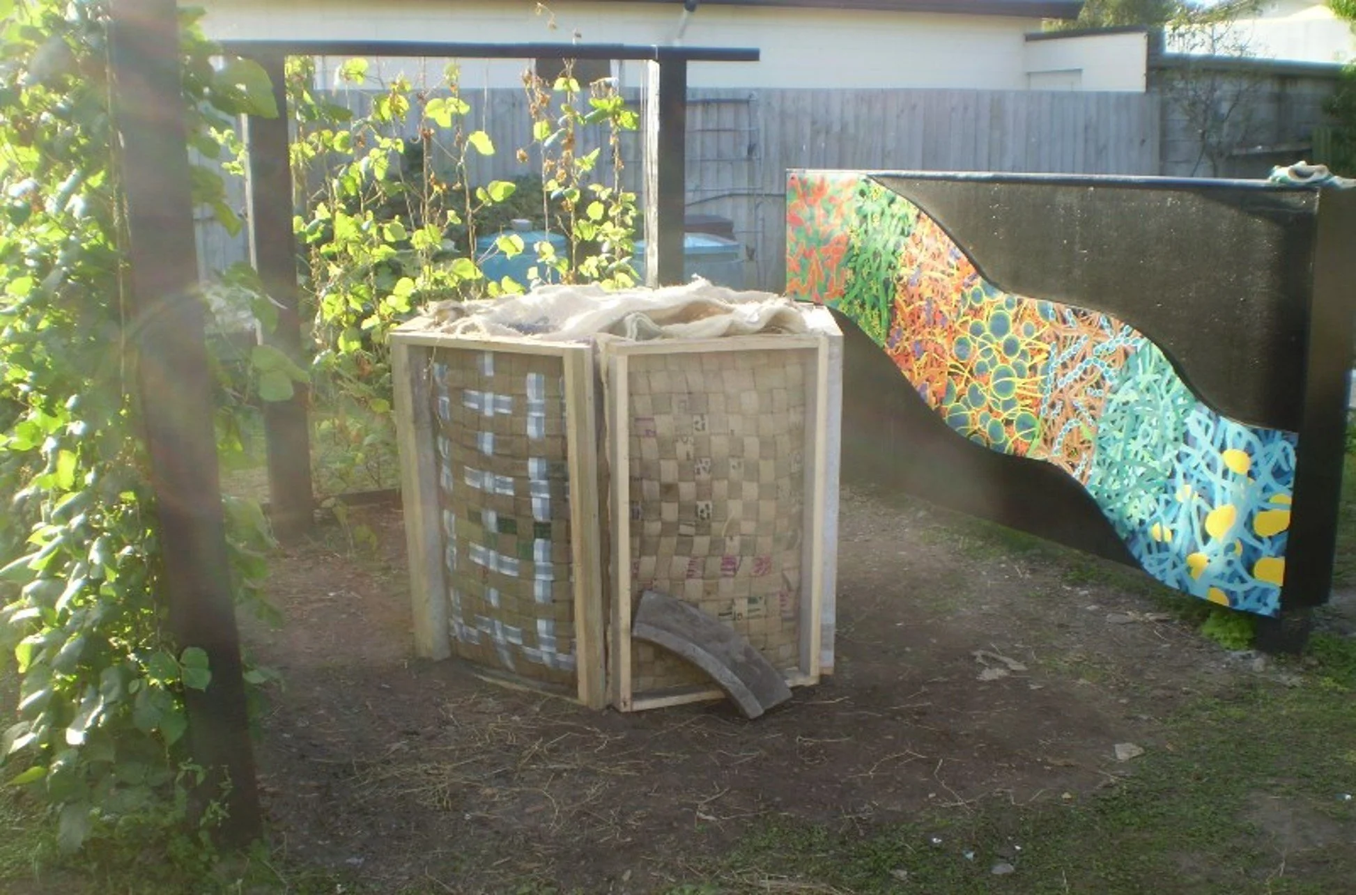 A compost bin made from wooden and woven materials in a backyard, with a colorful mural on a black fence in the background.