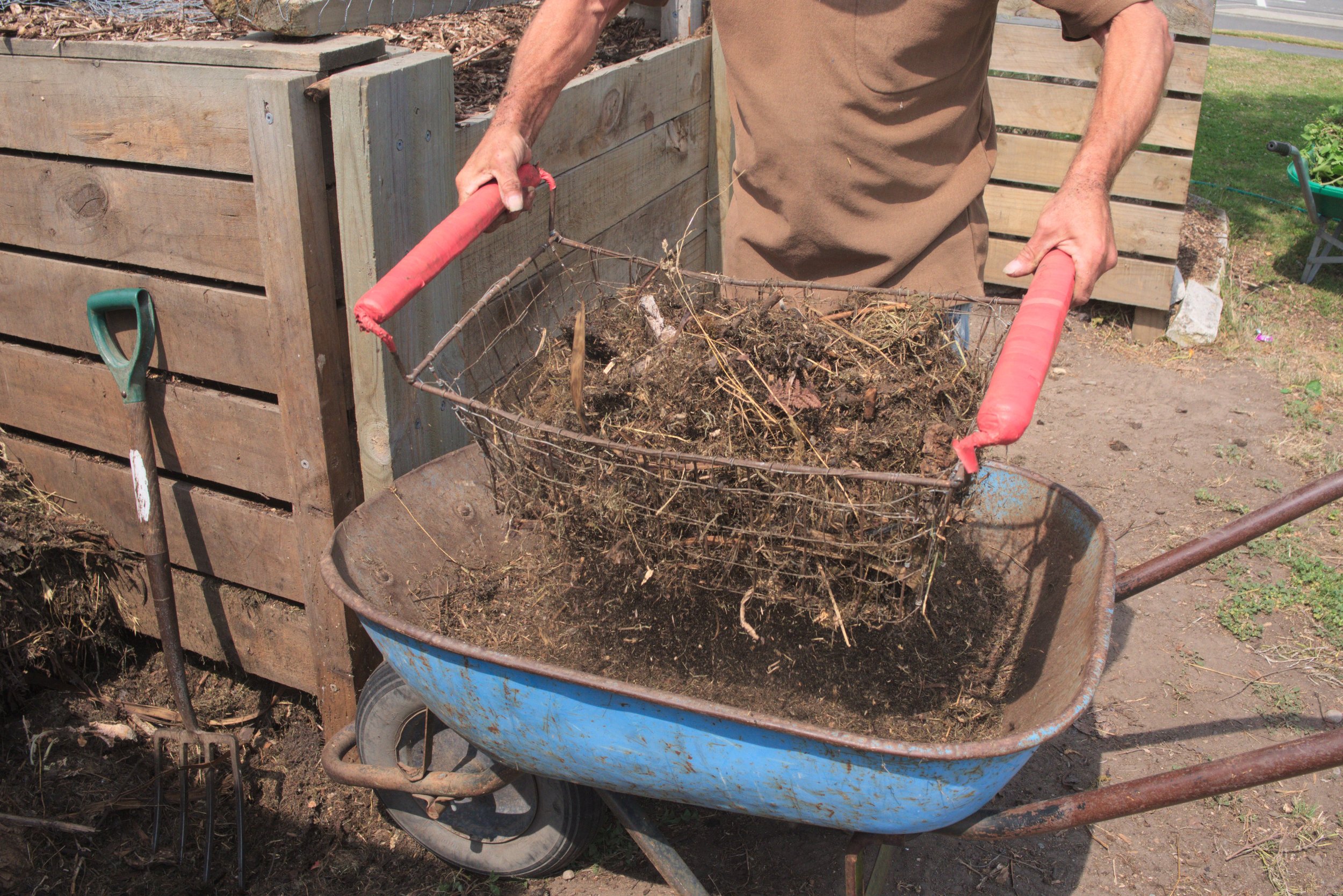 Person using a wire basket to remove dirt and debris from a wheelbarrow during gardening.