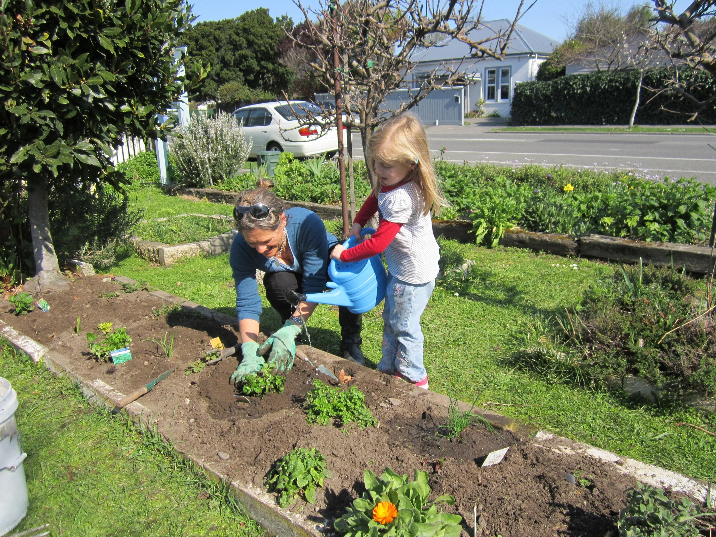 A woman and a young girl planting flowers or vegetables in a garden bed outdoors. The girl holds a watering can, and the woman is kneeling, tending the plants.