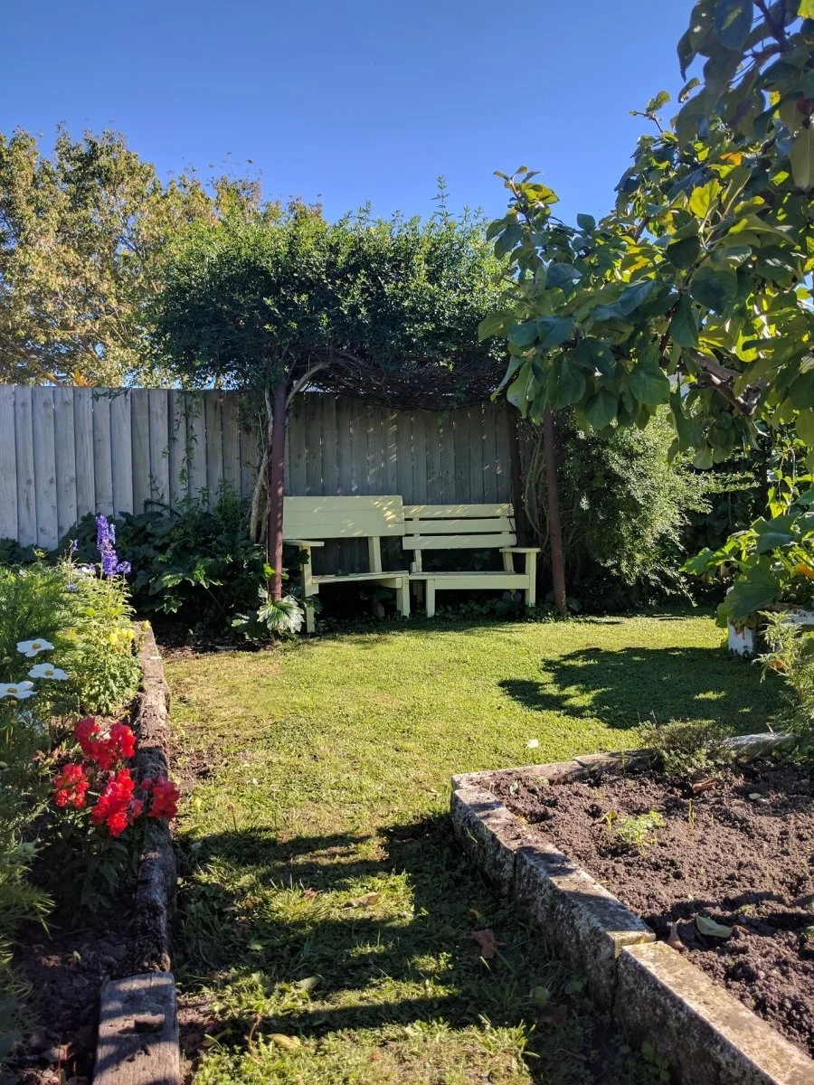 A garden with a wooden fence, vibrant flowers on the left, and a grassy area with a bench shade by trees on the back. Soil plot on the right.