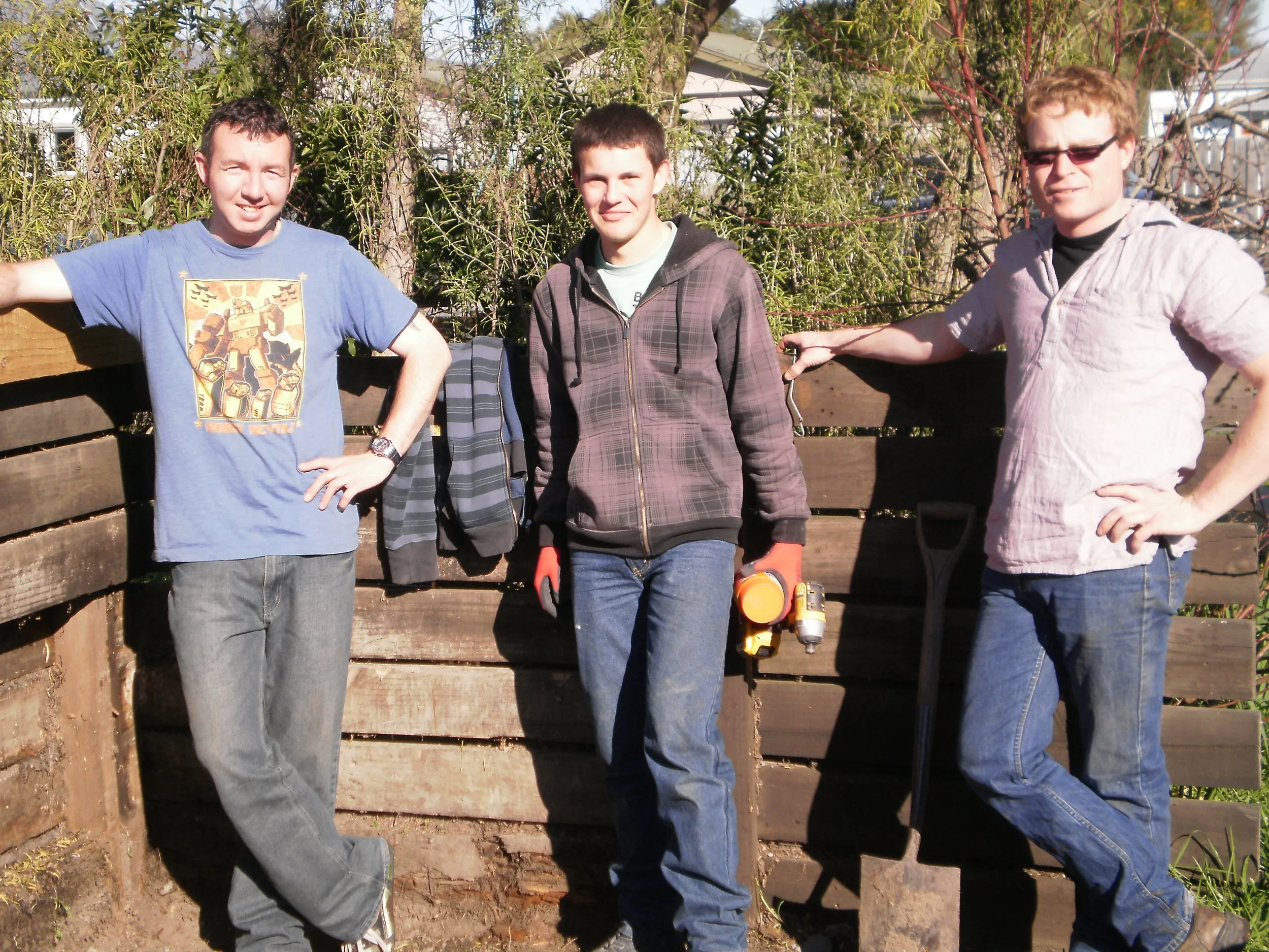 Three young men standing outdoors in front of a wooden fence, with trees in the background, posing for a photo. The man on the left is wearing a blue T-shirt with a graphic design, the man in the middle is wearing a gray hoodie with tools hanging from his pocket, and the man on the right is wearing a light-colored short-sleeve shirt and sunglasses.