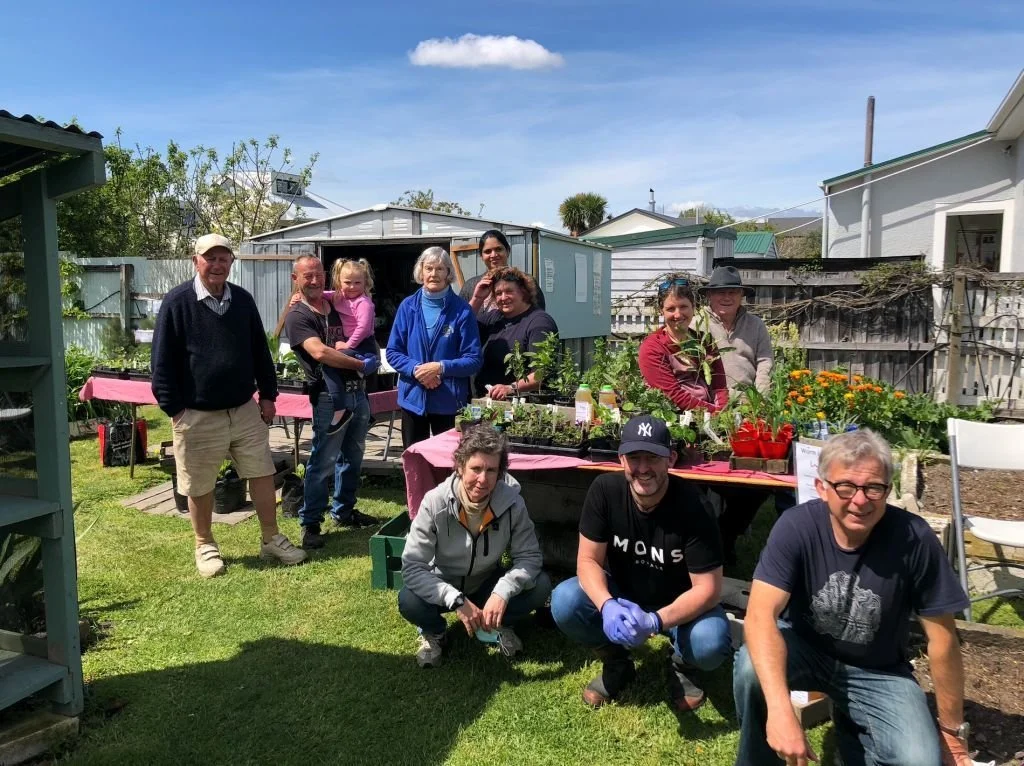 Group of people gathering outdoors in a garden, with tables of plants and flowers, in a residential backyard with sheds and houses in the background.