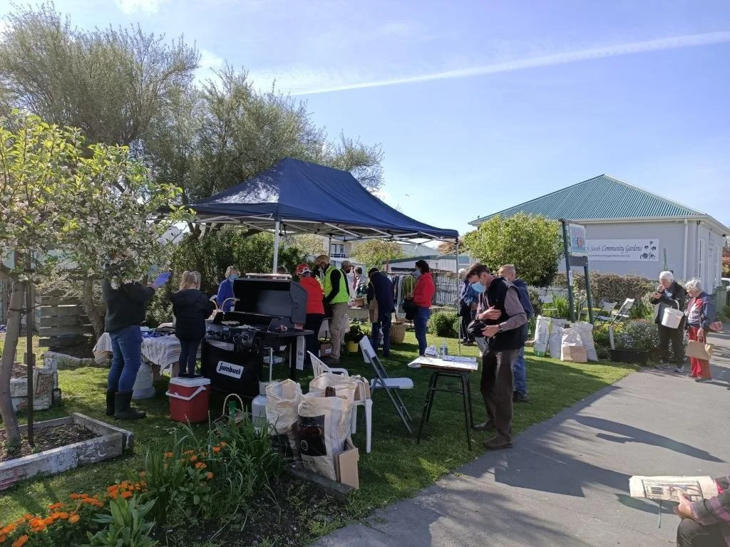 A community garden event with people gathered around a table under a blue canopy, some cooking on a grill, others browsing items or taking photos. There are flowering plants, trees, and a community center building in the background.
