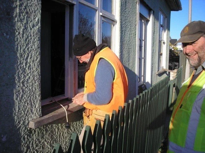 Two men working outside a residential building; one is inspecting or working near a window, the other is standing nearby smiling, both wearing safety vests and work hats.