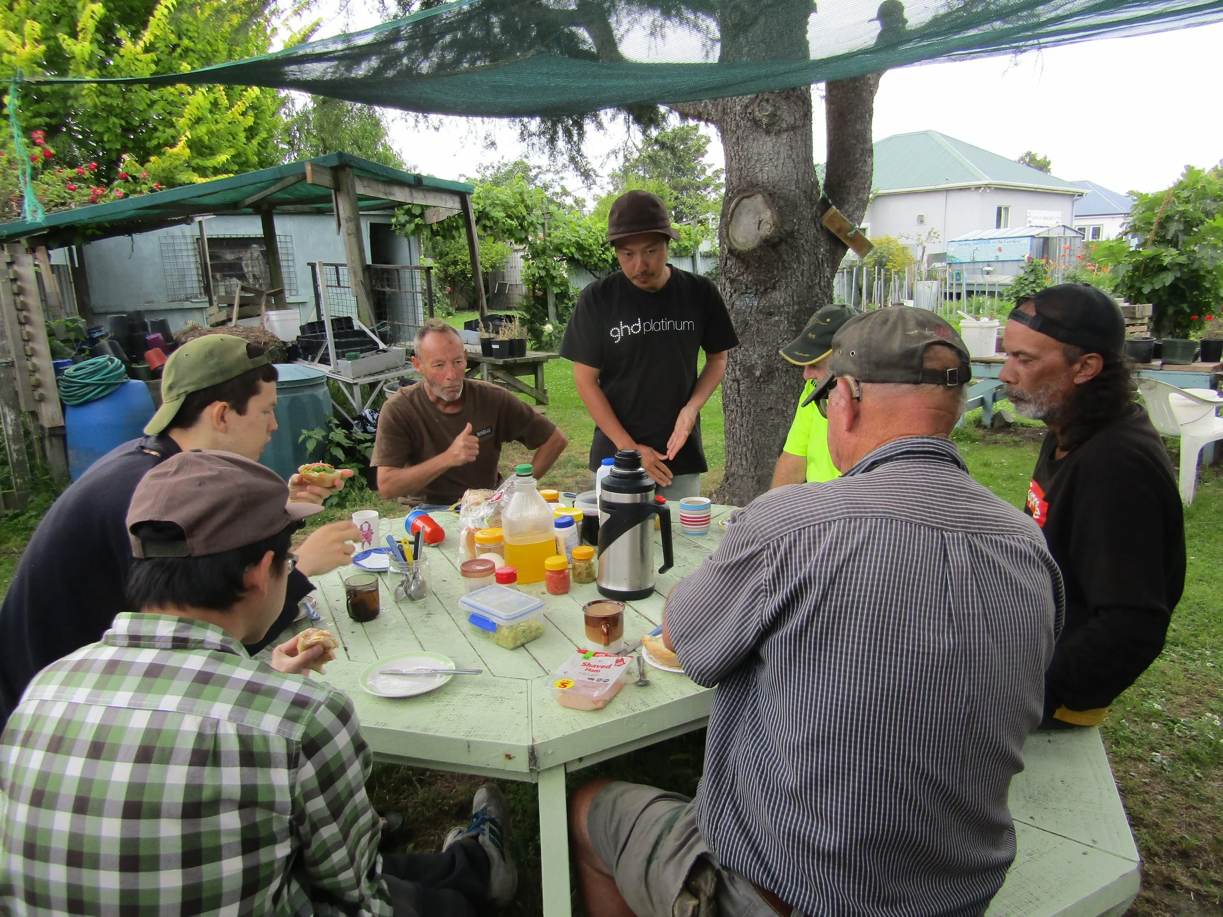 Group of people having a casual outdoor meal at a picnic table in a backyard, with food, drinks, and condiments on the table, surrounded by trees and garden structures.