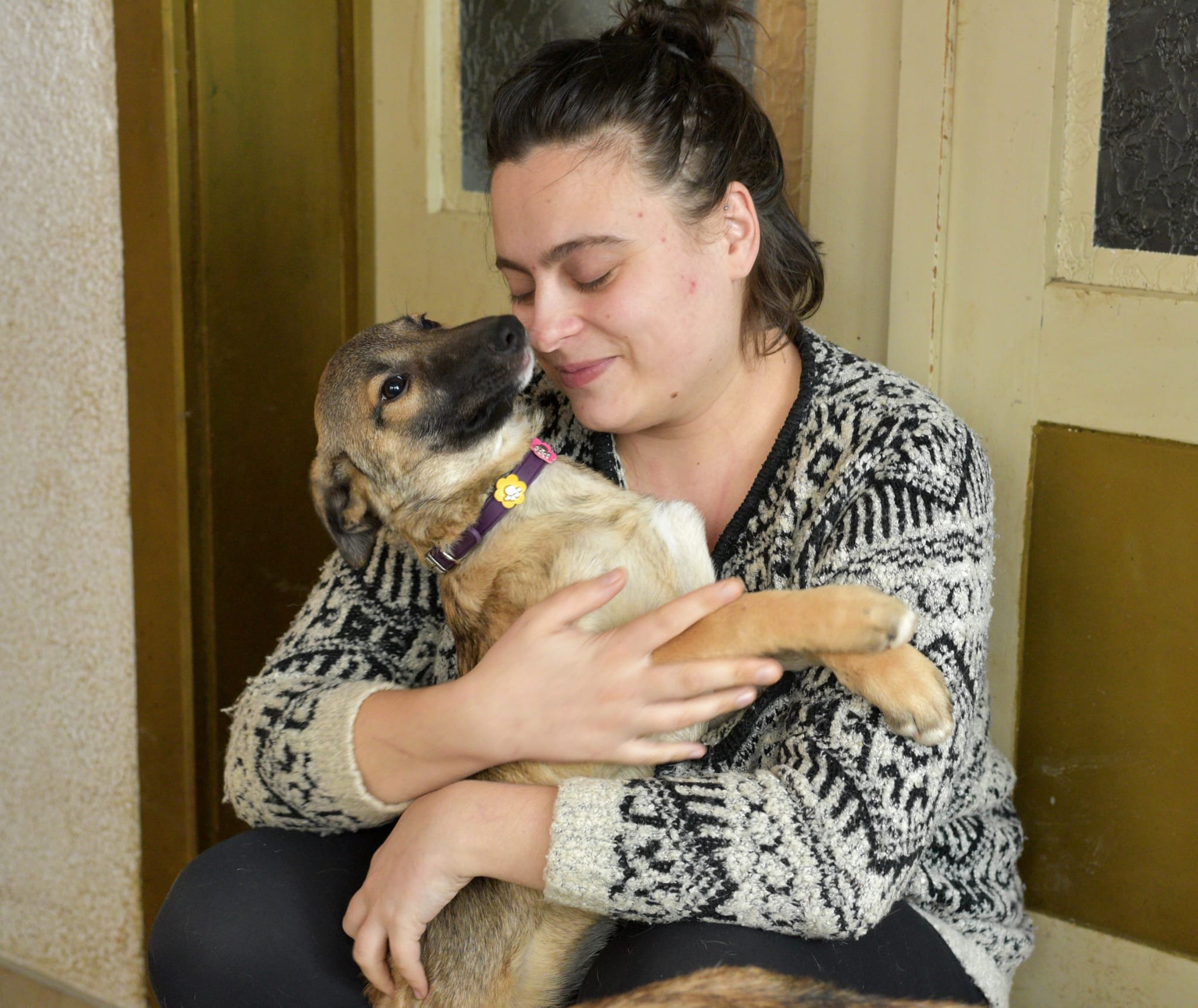 A young woman with dark hair in a bun, wearing a patterned sweater, holding a dog with light brown and black fur close to her face, smiling gently as the dog looks up at her.