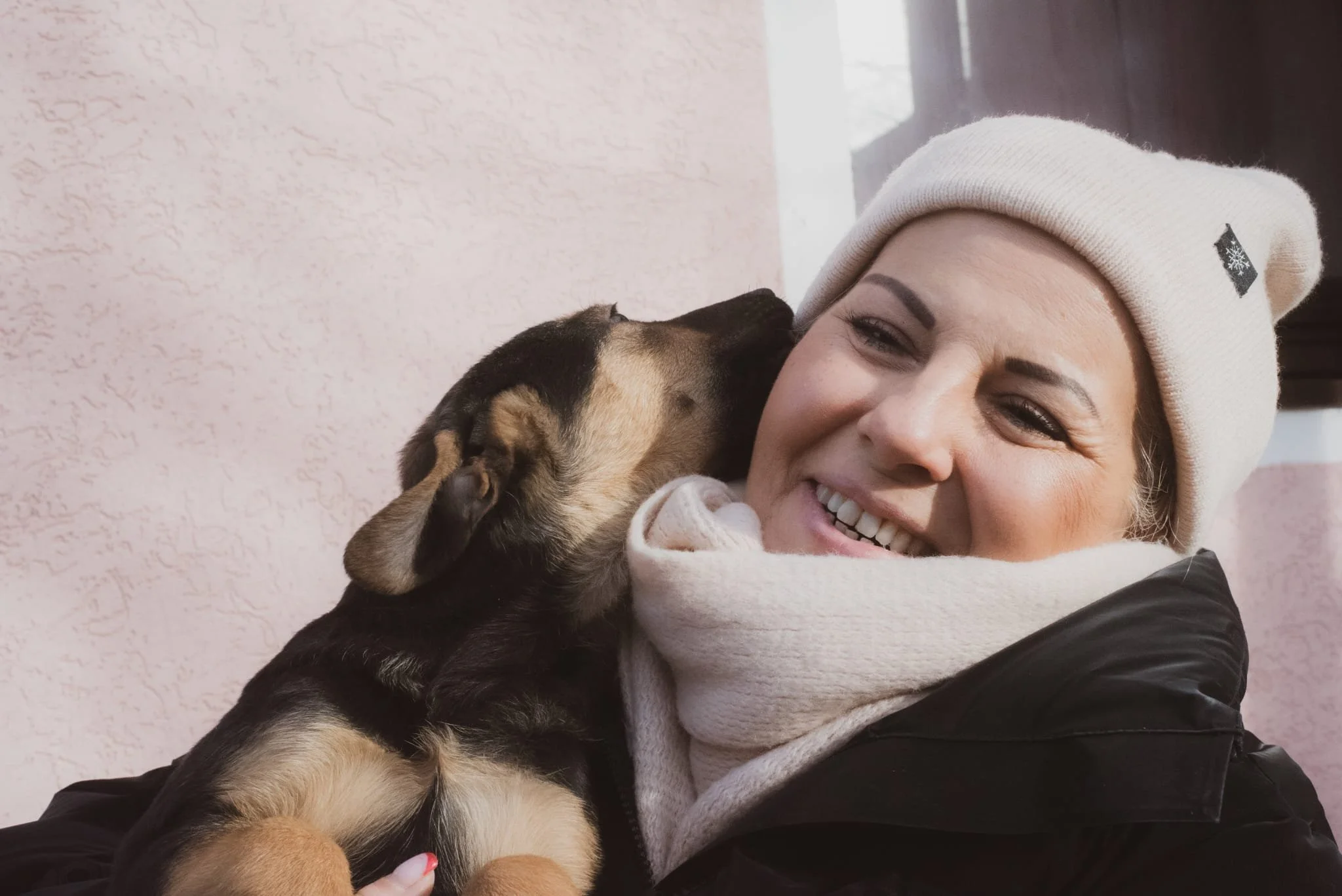 A woman smiling as a puppy licks her face, wearing winter clothing including a white beanie and scarf.