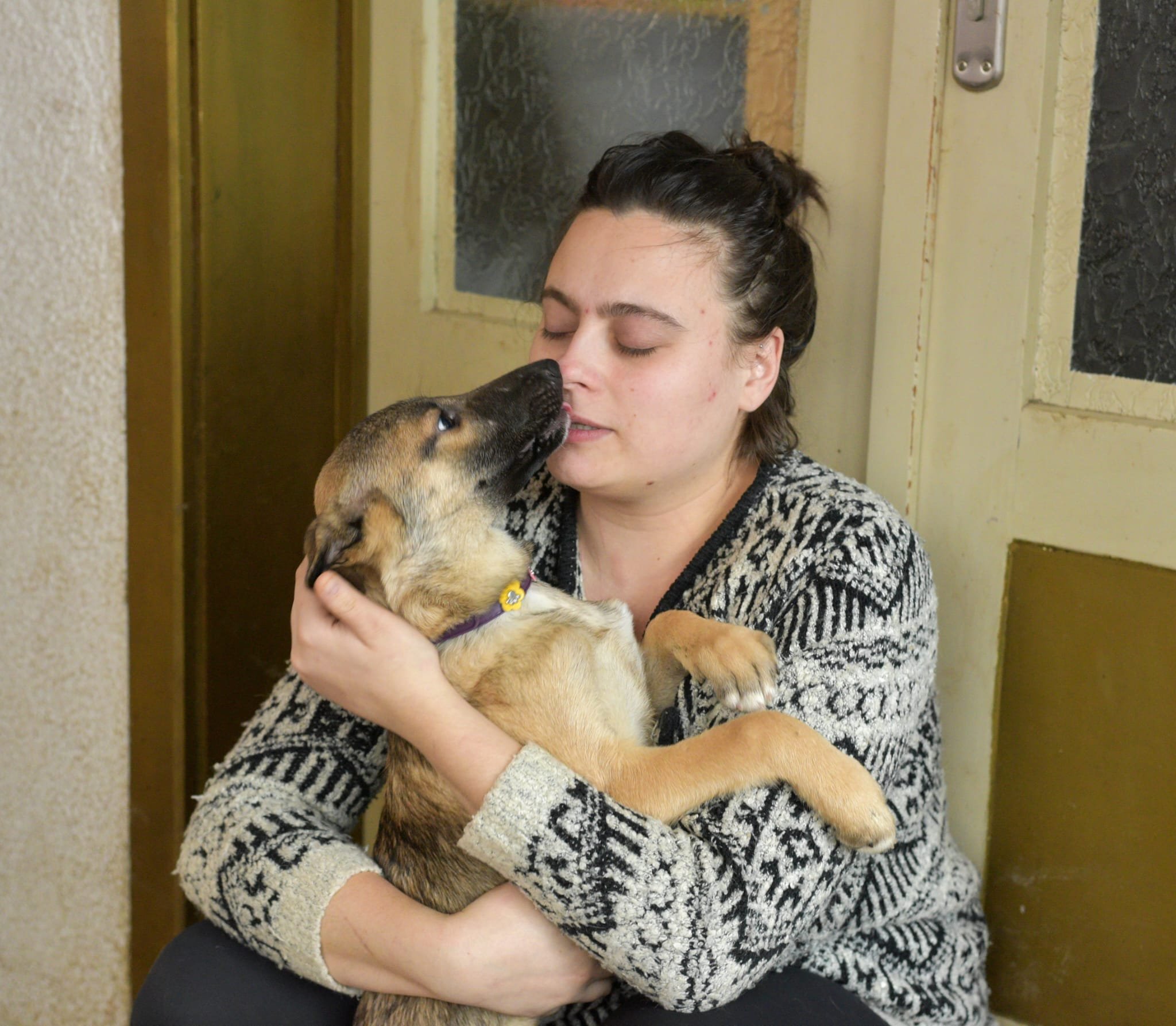 A woman with dark hair pulled back, wearing a patterned black and white sweater, holds a puppy close to her face while sitting indoors near a door.
