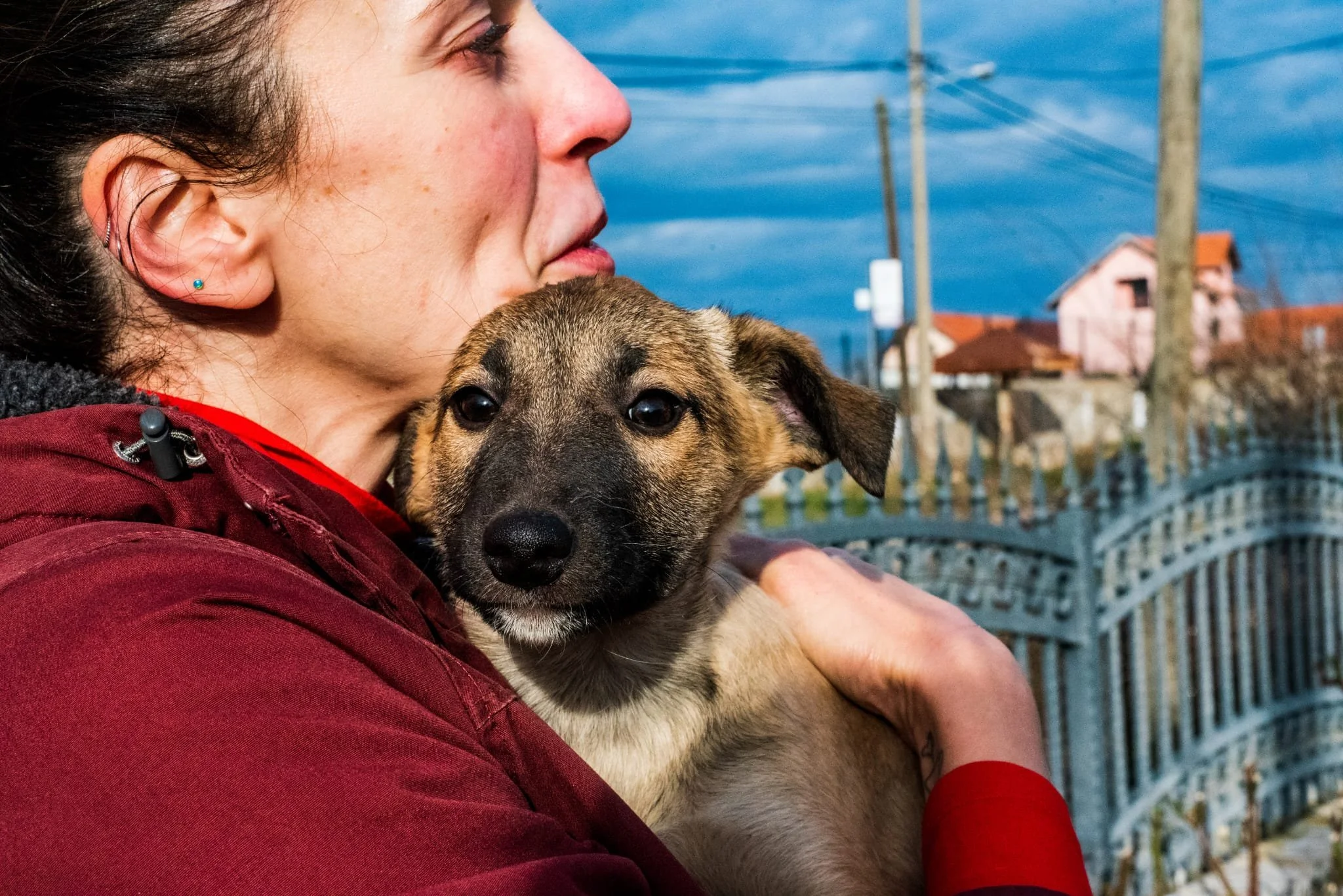 A woman holding a puppy close to her chest outdoors with a fence, utility poles, and houses in the background.