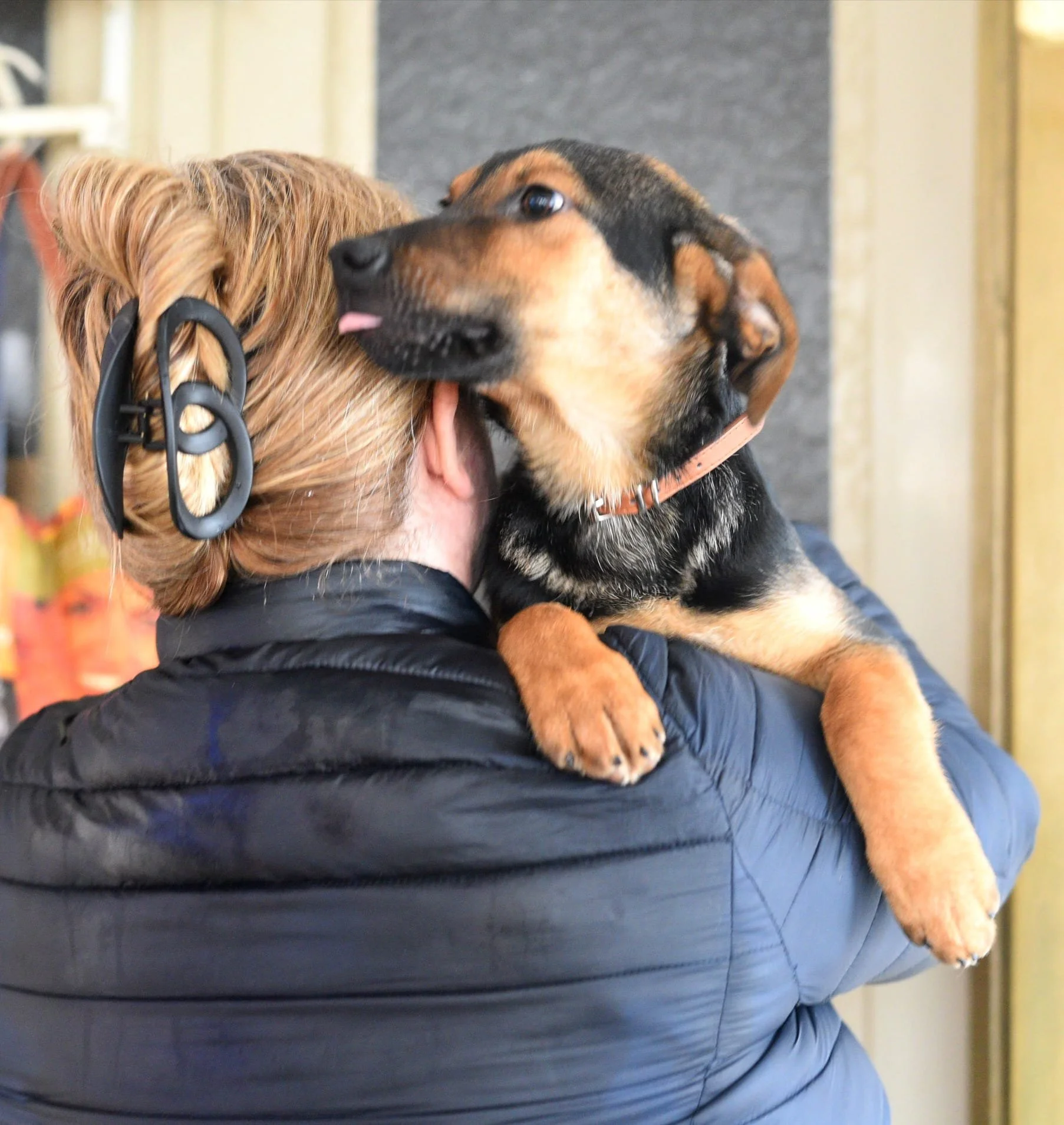 A person with red hair in a bun wearing a black jacket is holding a puppy. The puppy is brown and black with a collar and is resting its paws on the person's shoulder. The puppy is licking or sniffing the person's face.