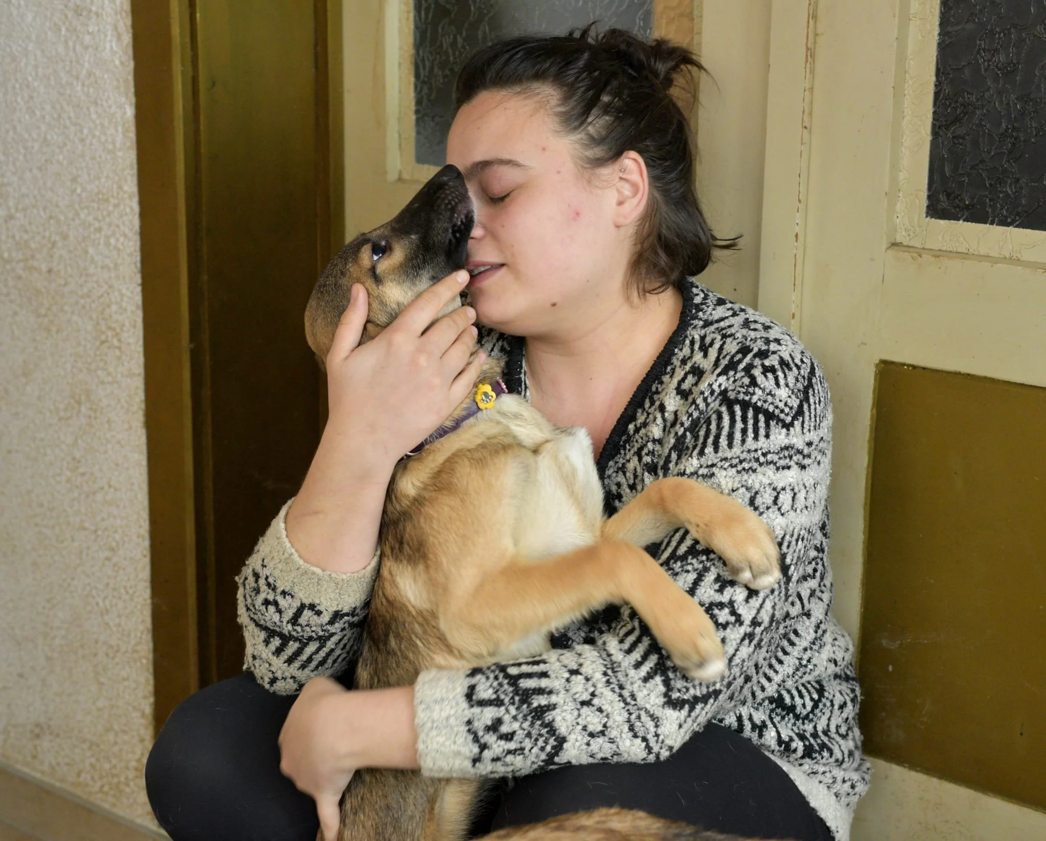 A woman with short dark hair, wearing a black and white patterned sweater, is sitting and holding a brown and black puppy close, gently touching noses with it in a cozy indoor setting.