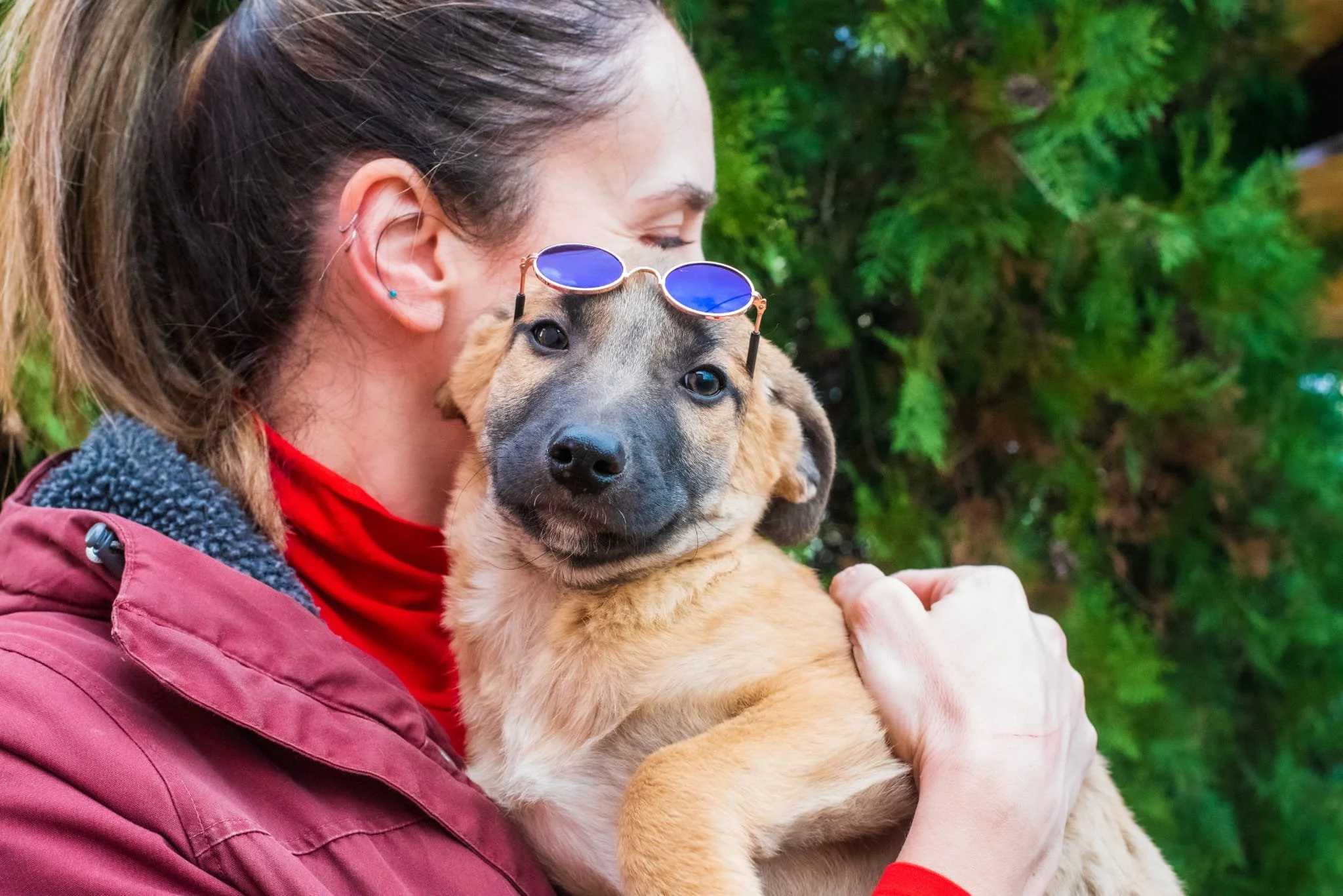 A woman with sunglasses on her head hugging a tan and black puppy outdoors, with green trees in the background.