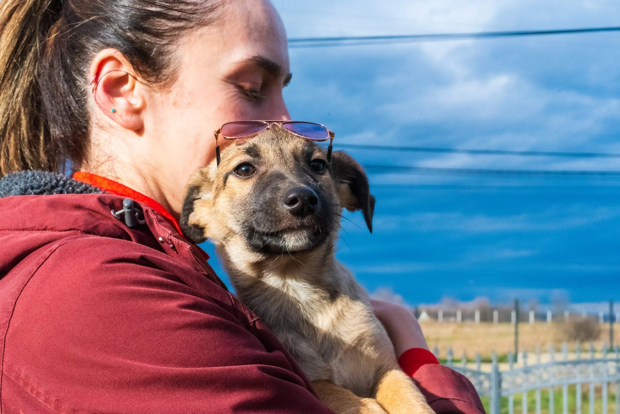 A young woman in a red jacket holding a puppy with sunglasses resting on its head outdoors, with a blue sky and landscape in the background.
