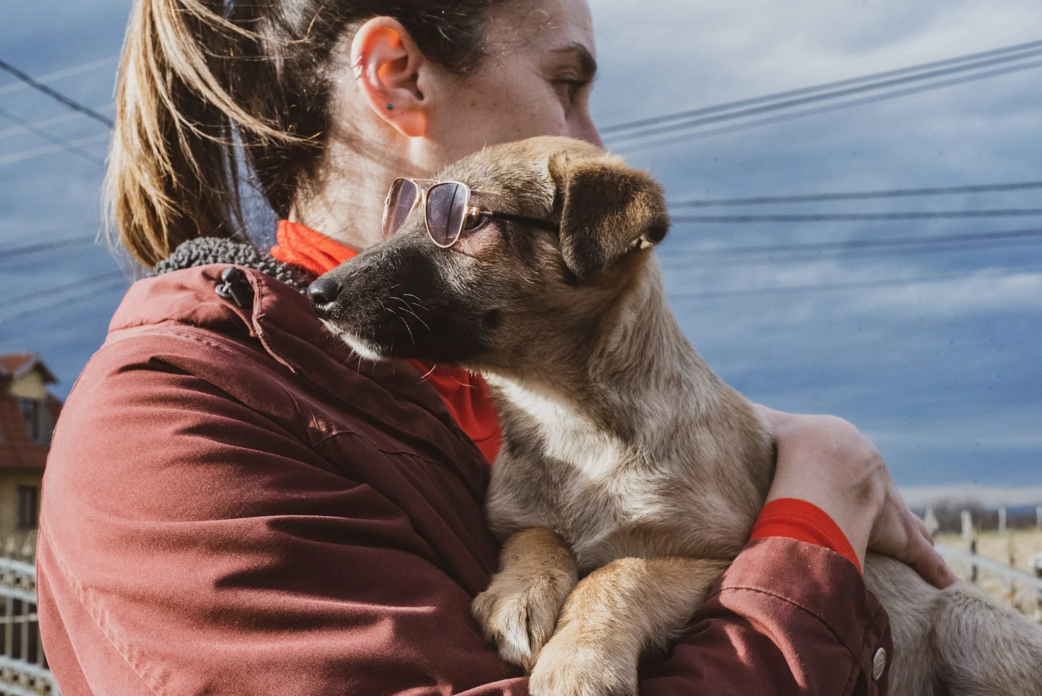 A woman holding a small dog wearing sunglasses outdoors on a cloudy day.
