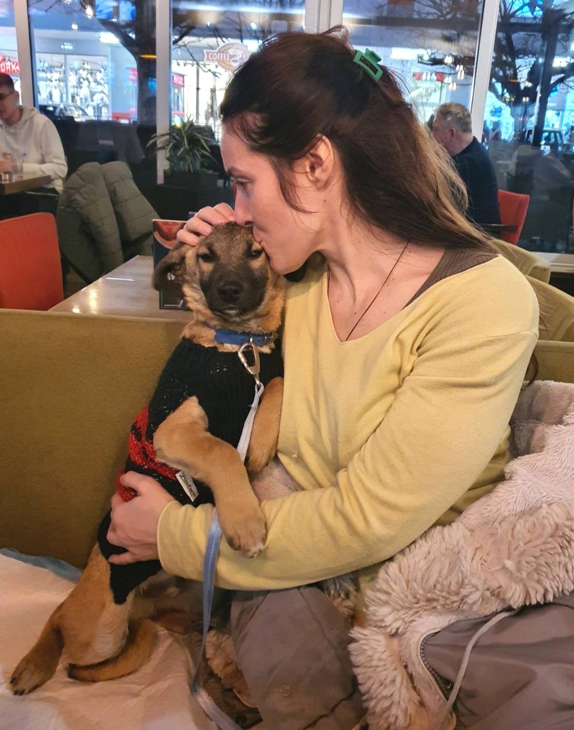 A woman with long dark hair and a green hair clip is holding a puppy and giving it a kiss on the head inside a cafe. The puppy is wearing a black sweater and a blue collar, sitting on her lap. The background shows other patrons and large windows.