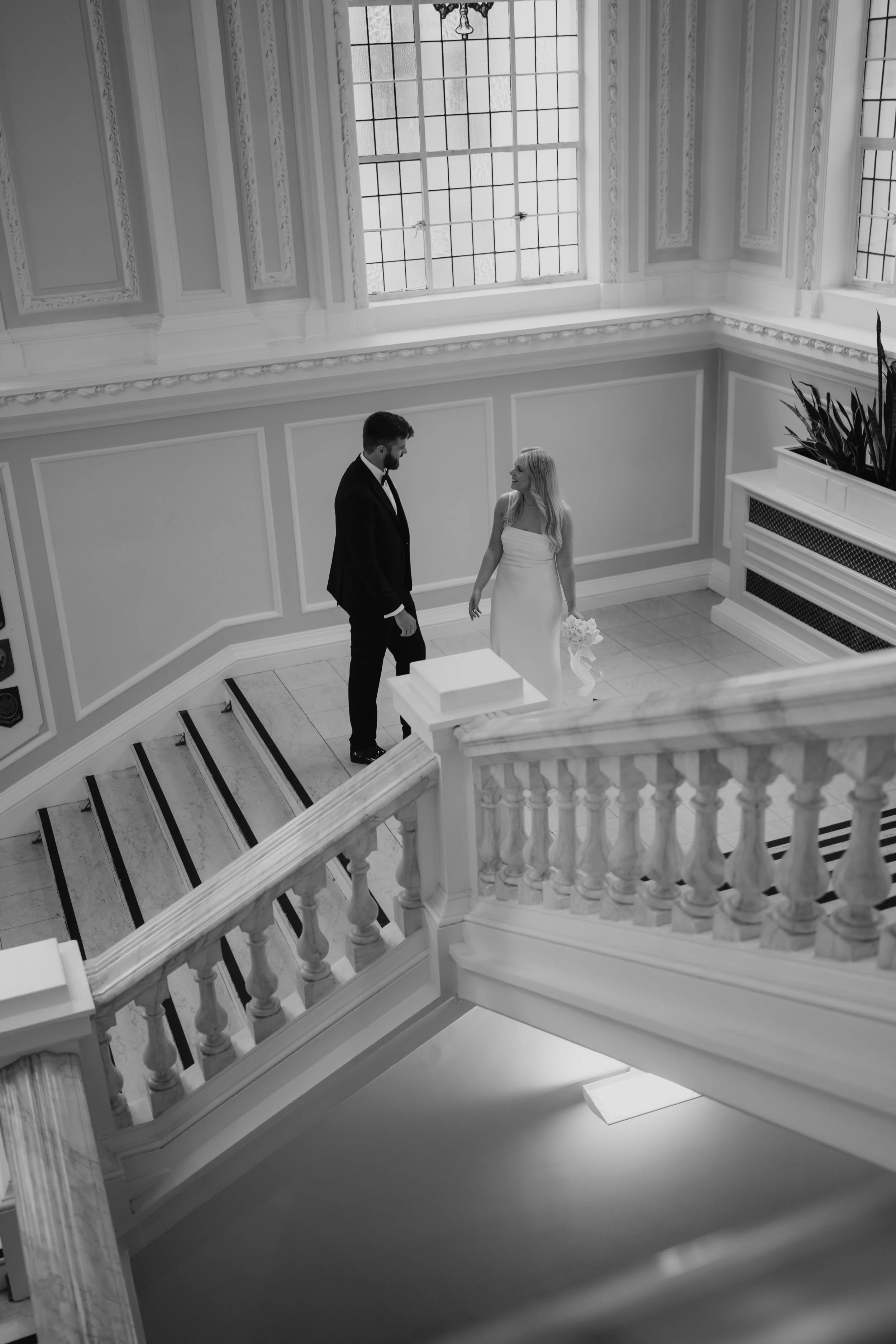 A couple dressed in formal attire in a bright, elegant hallway, standing near a staircase with a large window behind them.