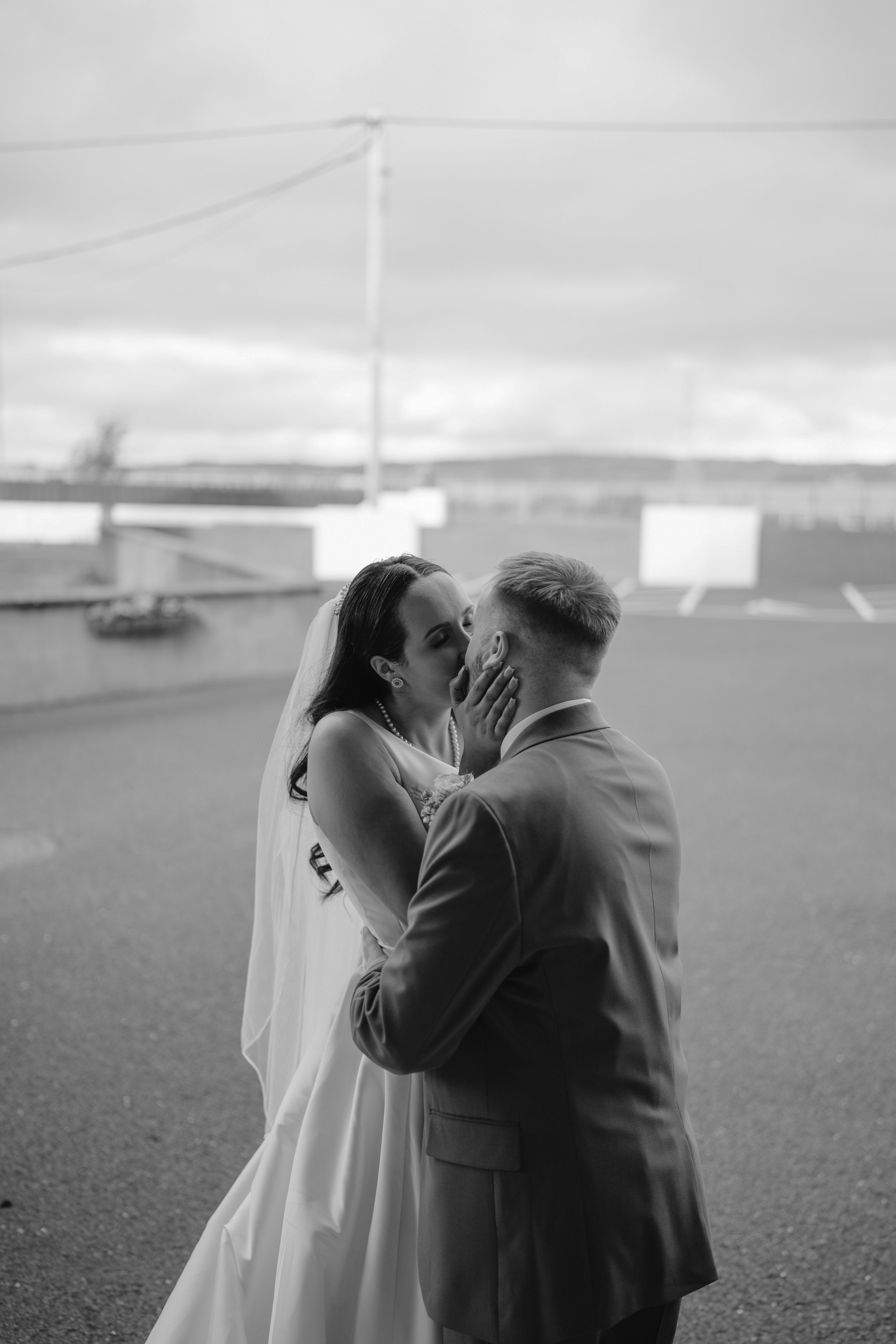 Black and white photo of a bride and groom sharing a kiss outdoors. The bride is wearing a wedding dress and veil, and the groom is in a suit. The bride is gently holding the groom's face with her hands.