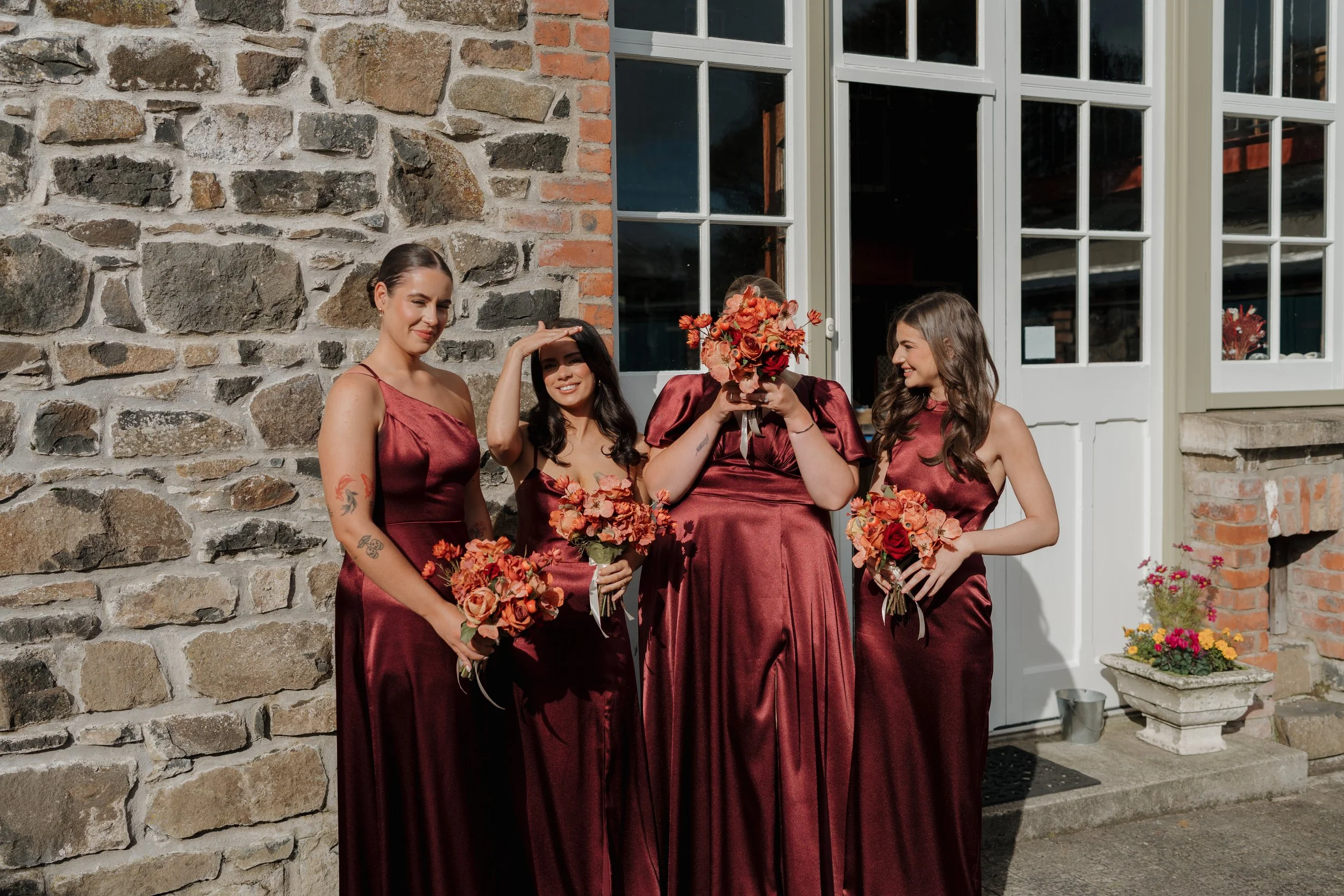Four women dressed in matching burgundy bridesmaid dresses standing outside in front of a stone wall and window, with one holding a bouquet of flowers, smiling and posing for the photo.