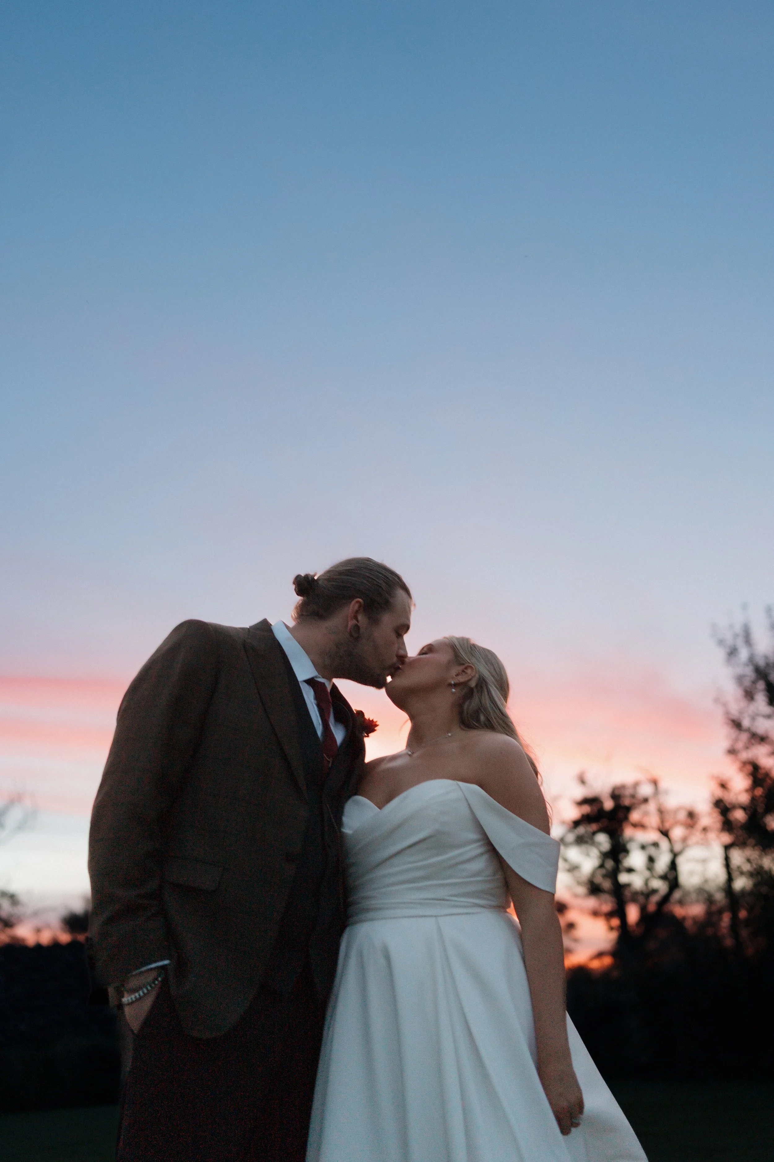 A bride and groom sharing a kiss during sunset, outdoors with silhouettes of trees in the background.