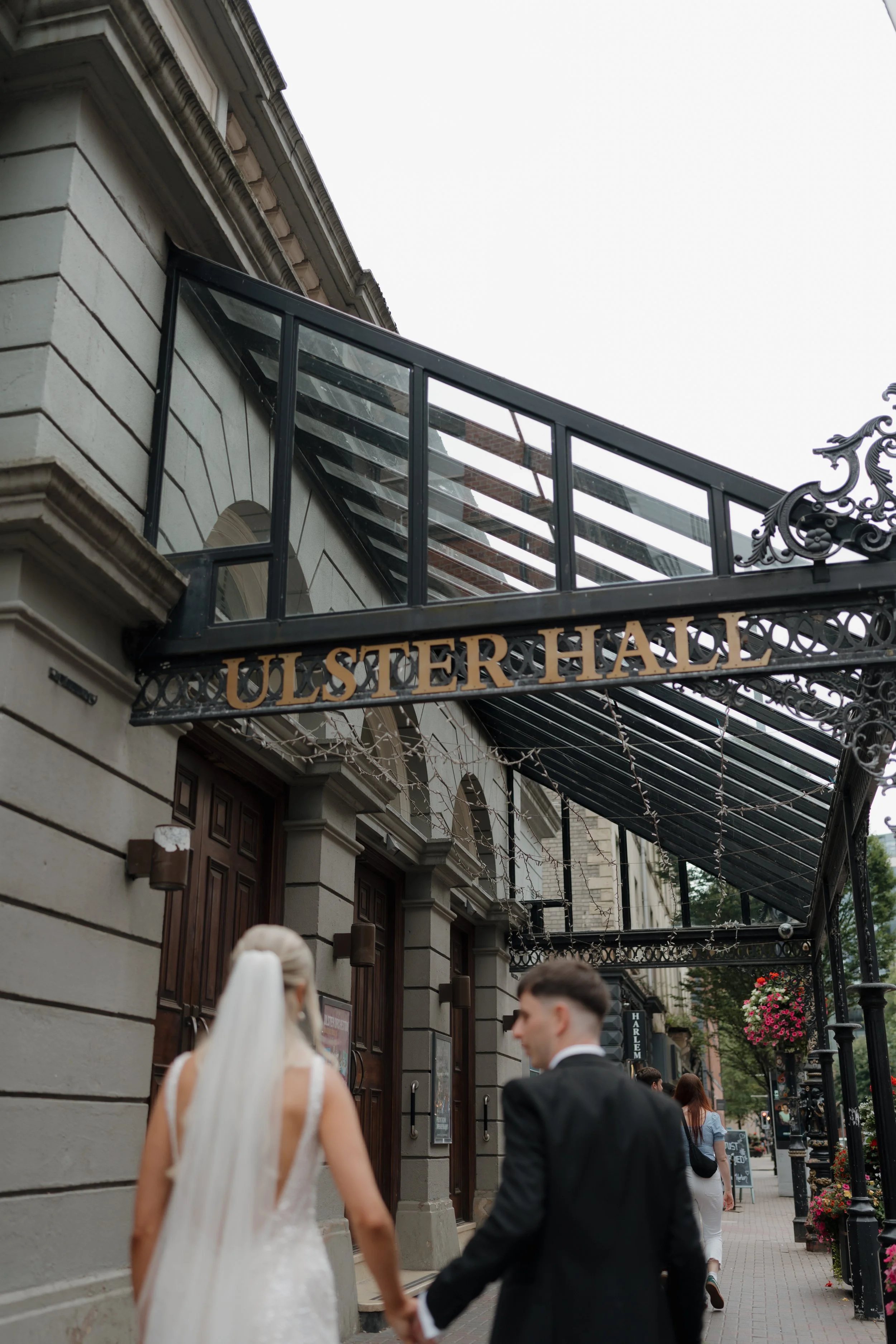 A bride and groom holding hands walk past the sign for Ulster Hall, a historic building with ornate metalwork on the exterior, decorative stone details, and string lights hanging above the sidewalk, in an urban setting with flowers and other pedestrians.