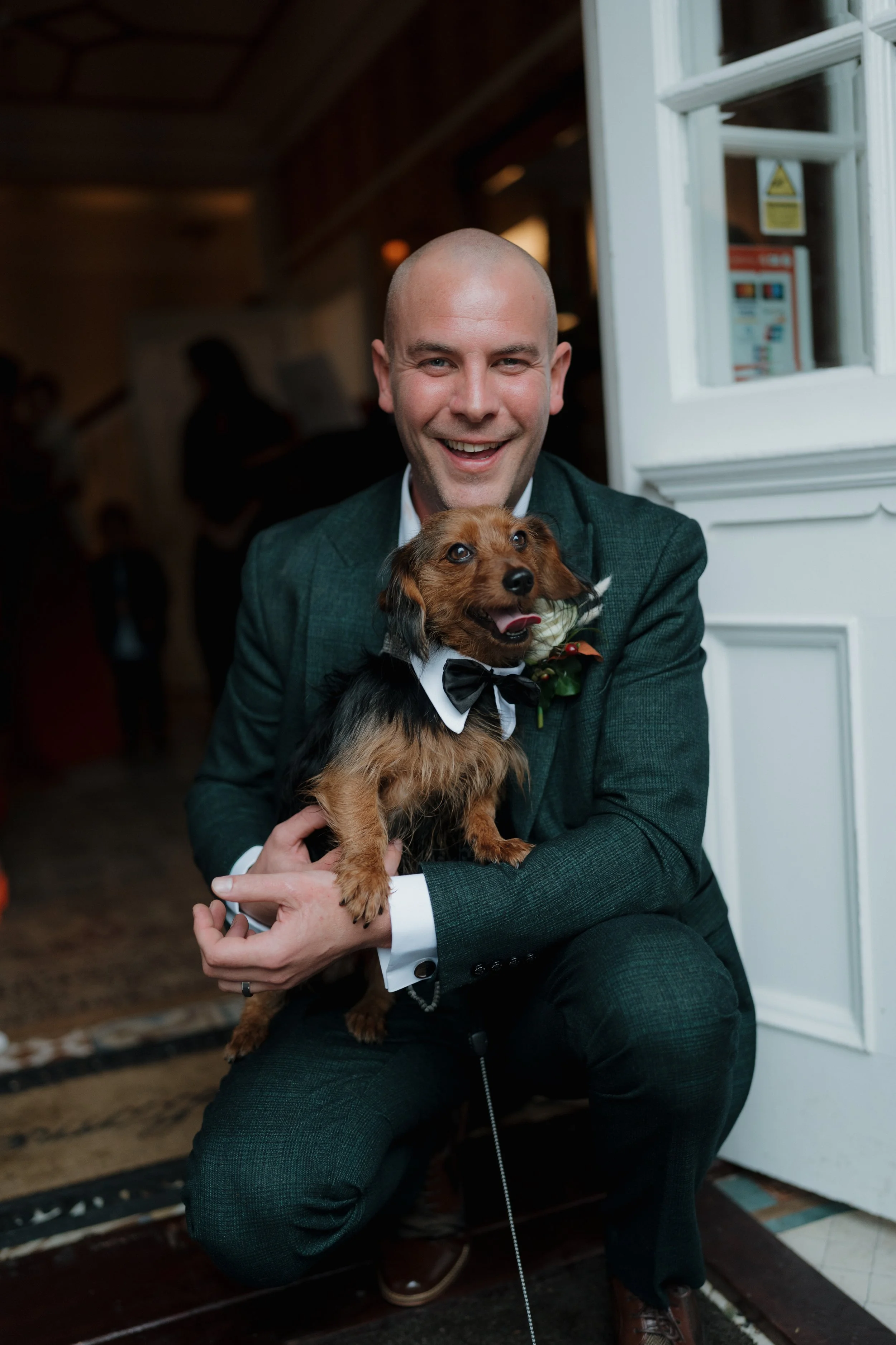A smiling man in a green suit holding a small dog dressed in a tuxedo with a bow tie at an indoor event.
