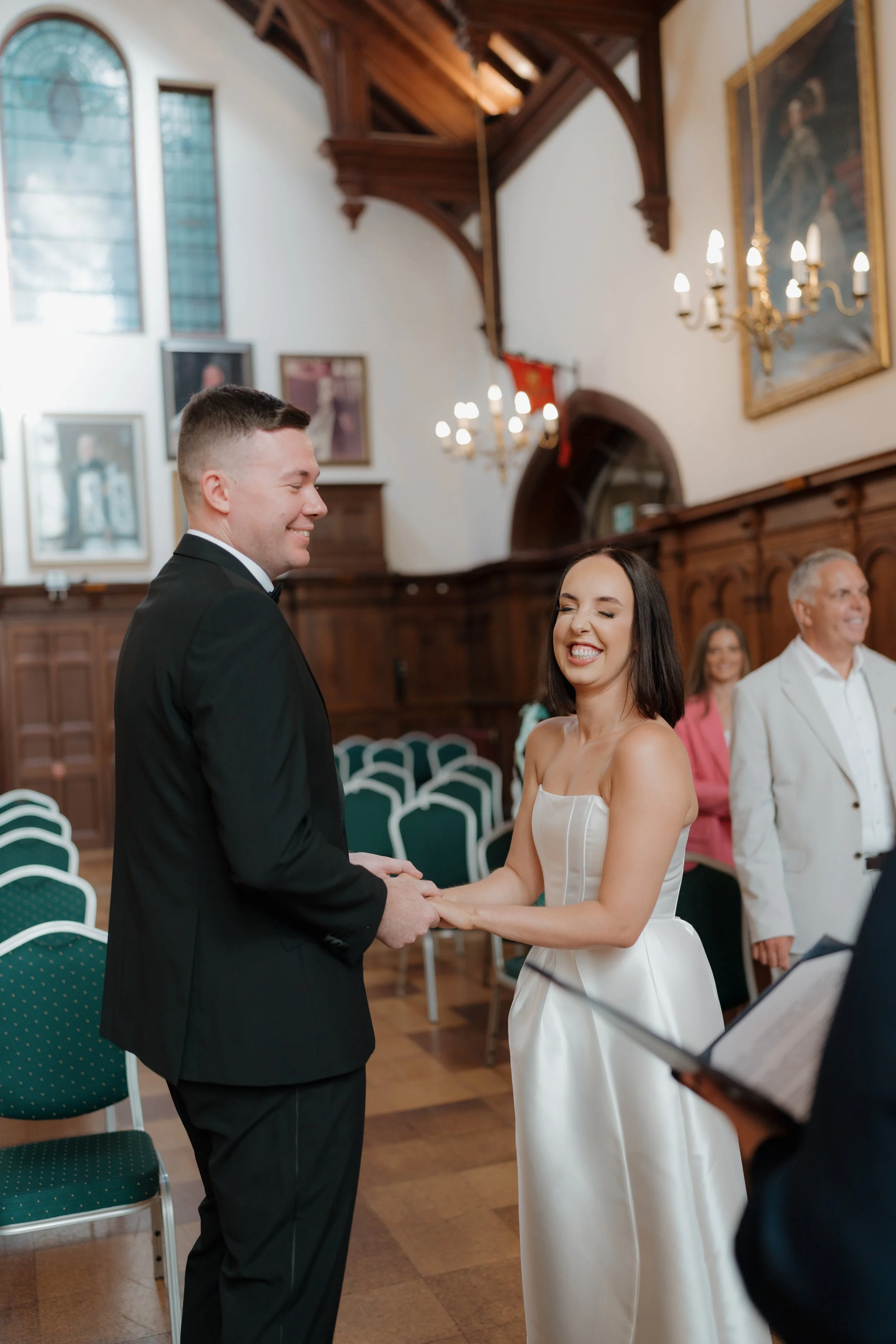 A couple marrying in a church, holding hands and smiling. The bride is wearing a strapless white wedding gown and the groom a black tuxedo. Guests are seated in the background.