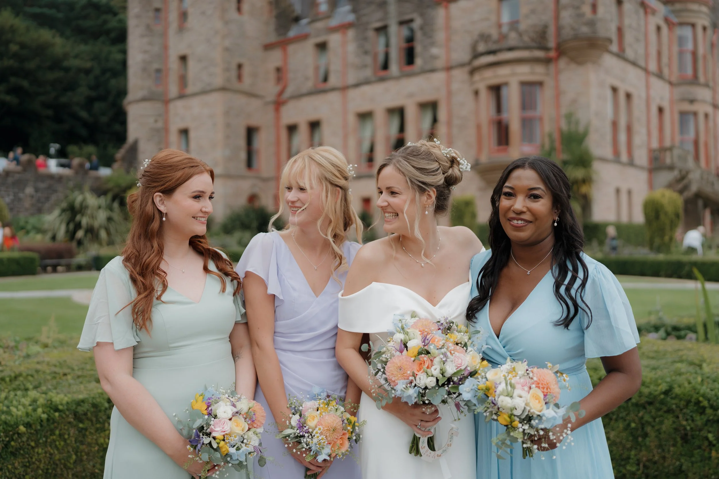 Group of four women in formal dresses standing outdoors on a wedding day, holding bouquets of flowers, with a large building and greenery in the background.
