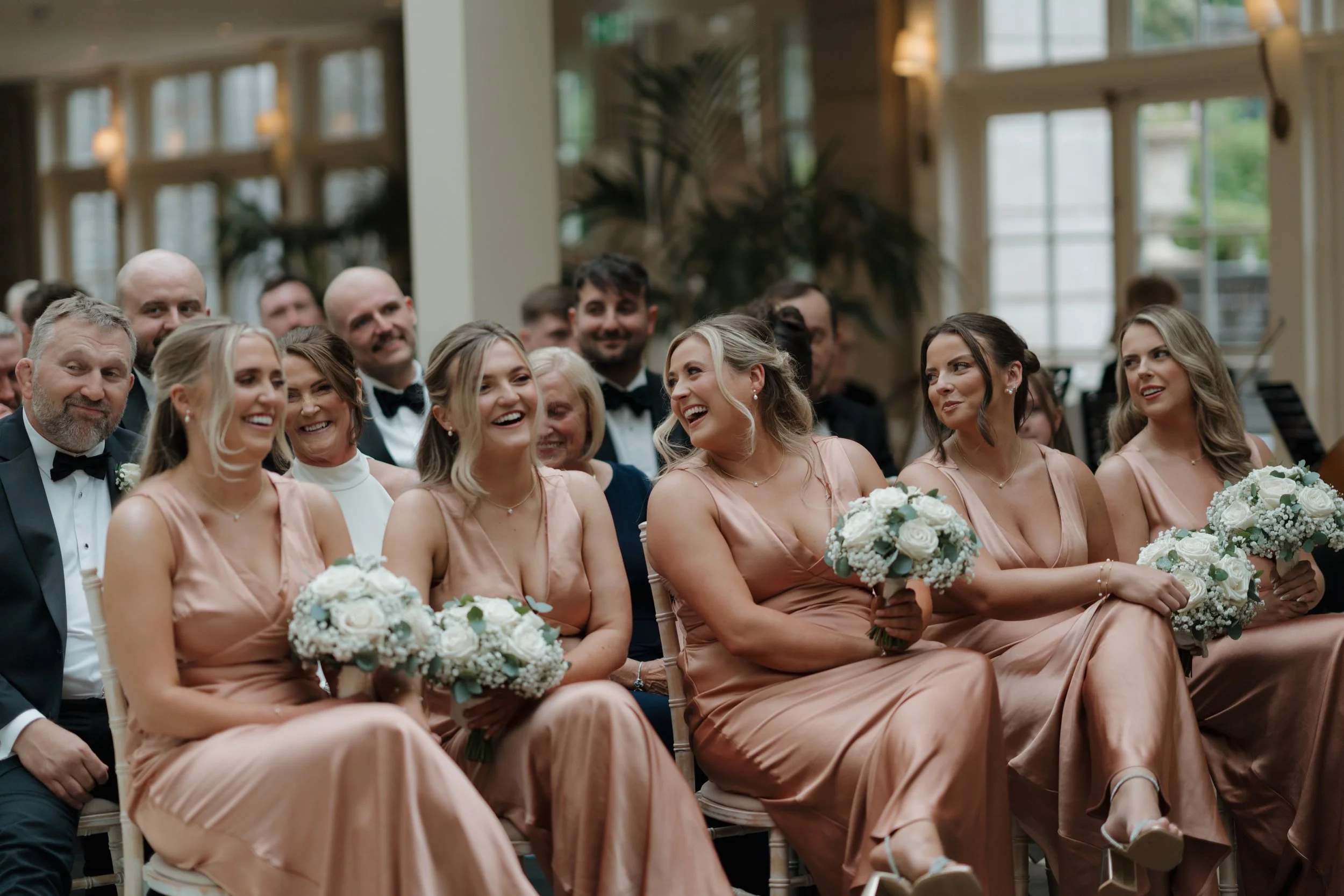 A group of wedding guests, including bridesmaids in peach-colored dresses holding bouquets, seated in an indoor venue, smiling and laughing during a wedding ceremony.