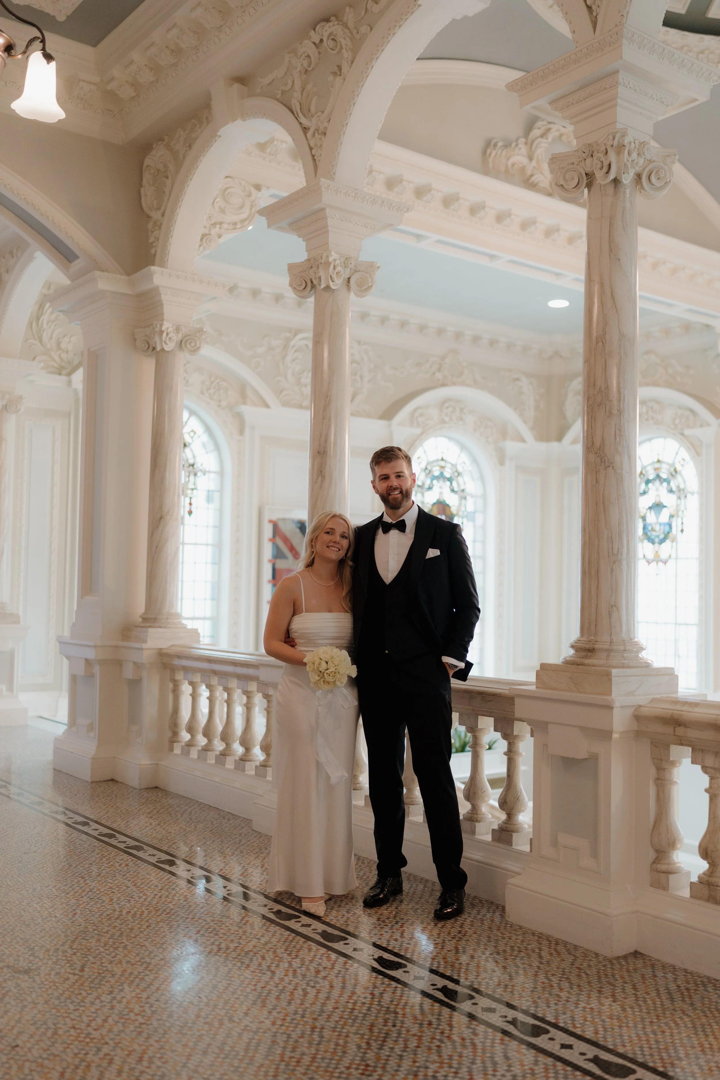 A bride and groom stand inside a grand, ornate building with tall marble columns, stained glass windows, and decorative architectural details. The bride is wearing a white wedding dress, and the groom is in a black tuxedo. The bride is holding a bouq