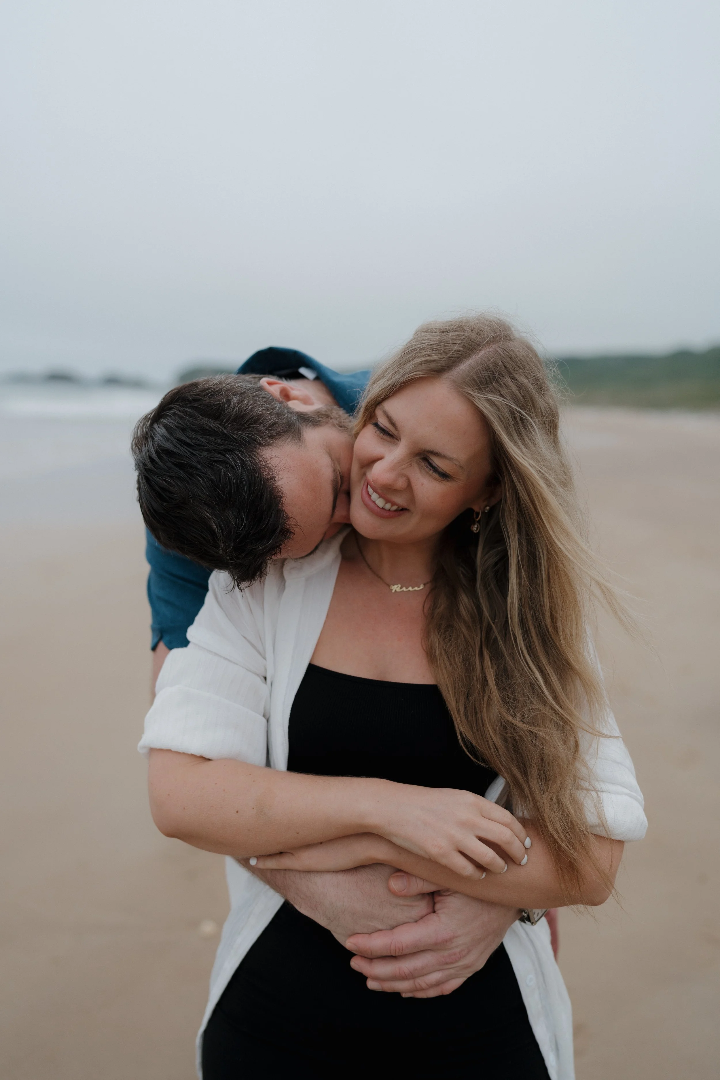 A couple on the beach, with the man kissing the woman's cheek from behind, and the woman smiling while looking down.