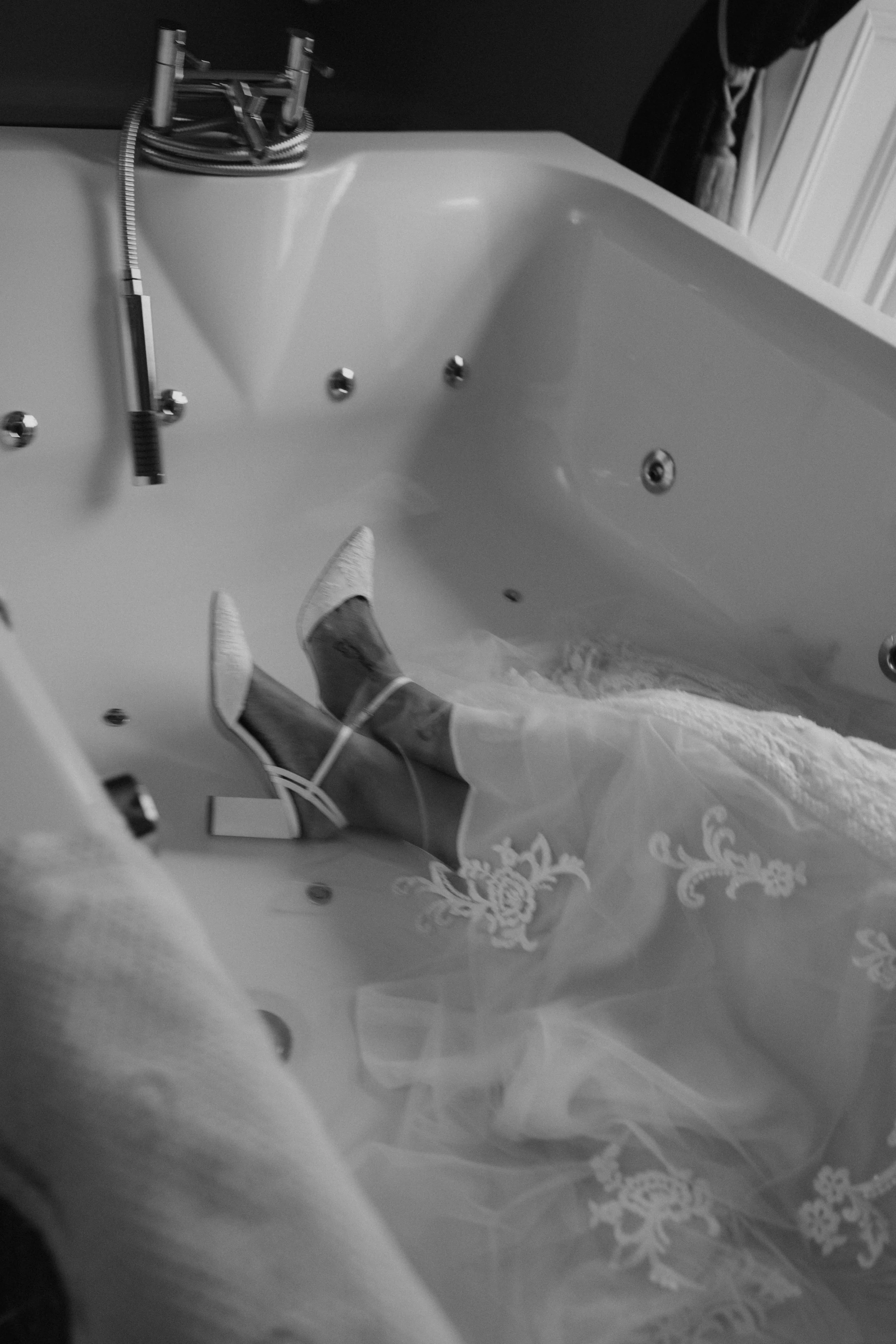 A person in a wedding dress lying in a bathtub with shoes on, surrounded by wedding veil and lace fabric.