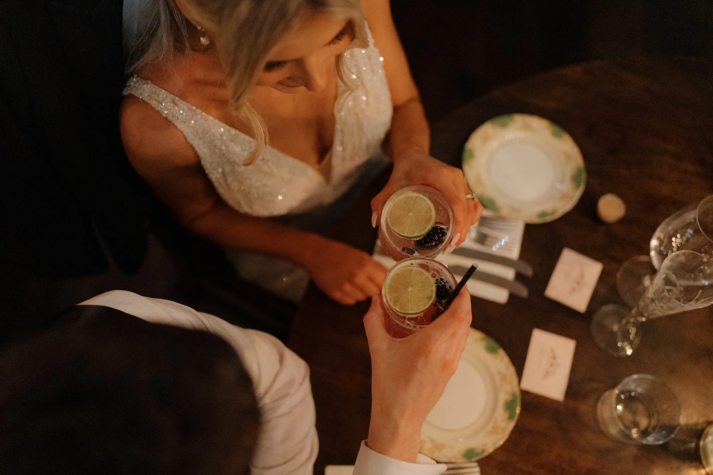 Bride and groom in formal dress holding glasses of cocktails with lemon slices and blackberries, seen from above at a dinner table with plates, napkins, and champagne glasses at Harlem Cafe Belfast.