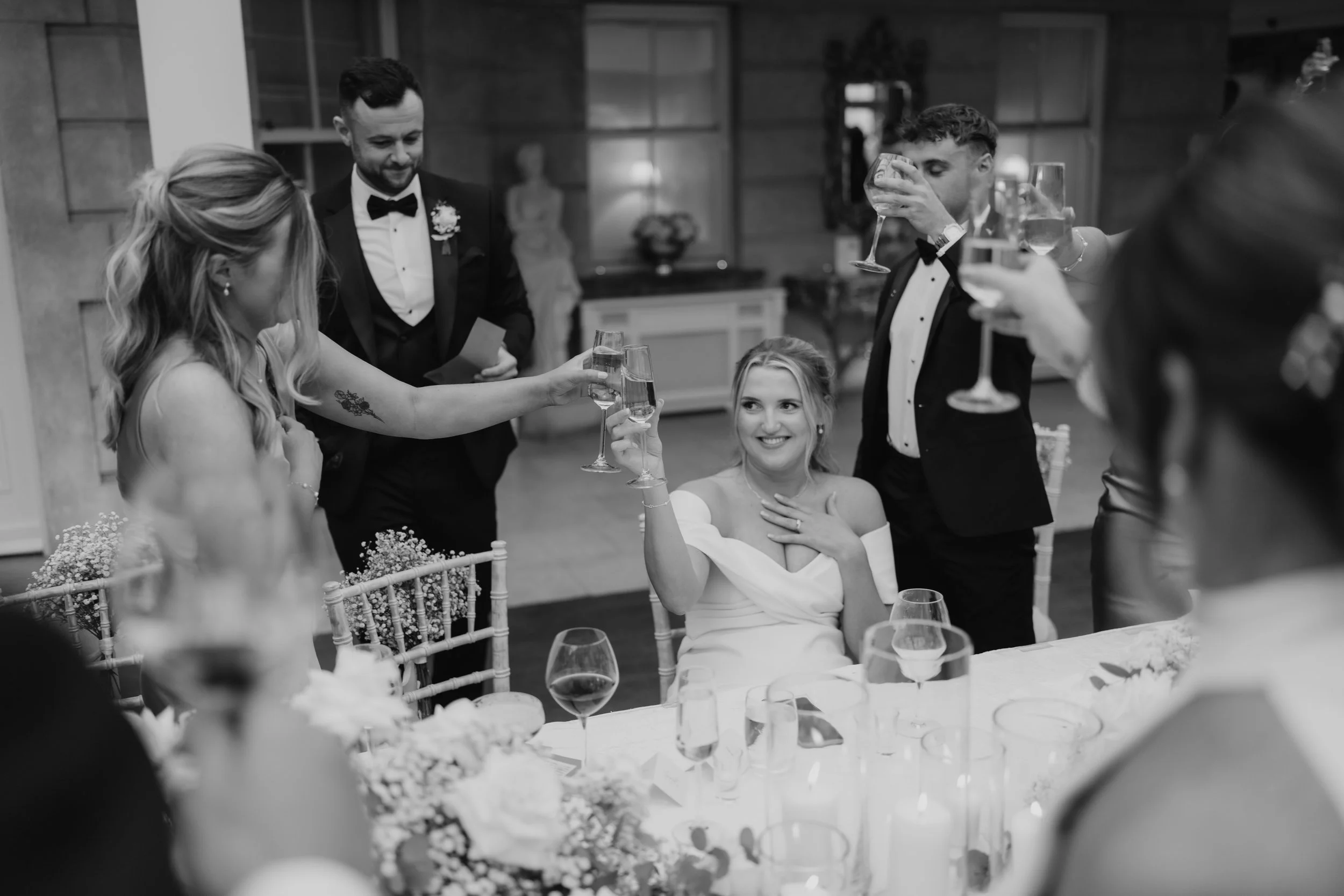People at a wedding celebration raising glasses for a toast, with a bride sitting at the table smiling and holding a glass, surrounded by friends and family.