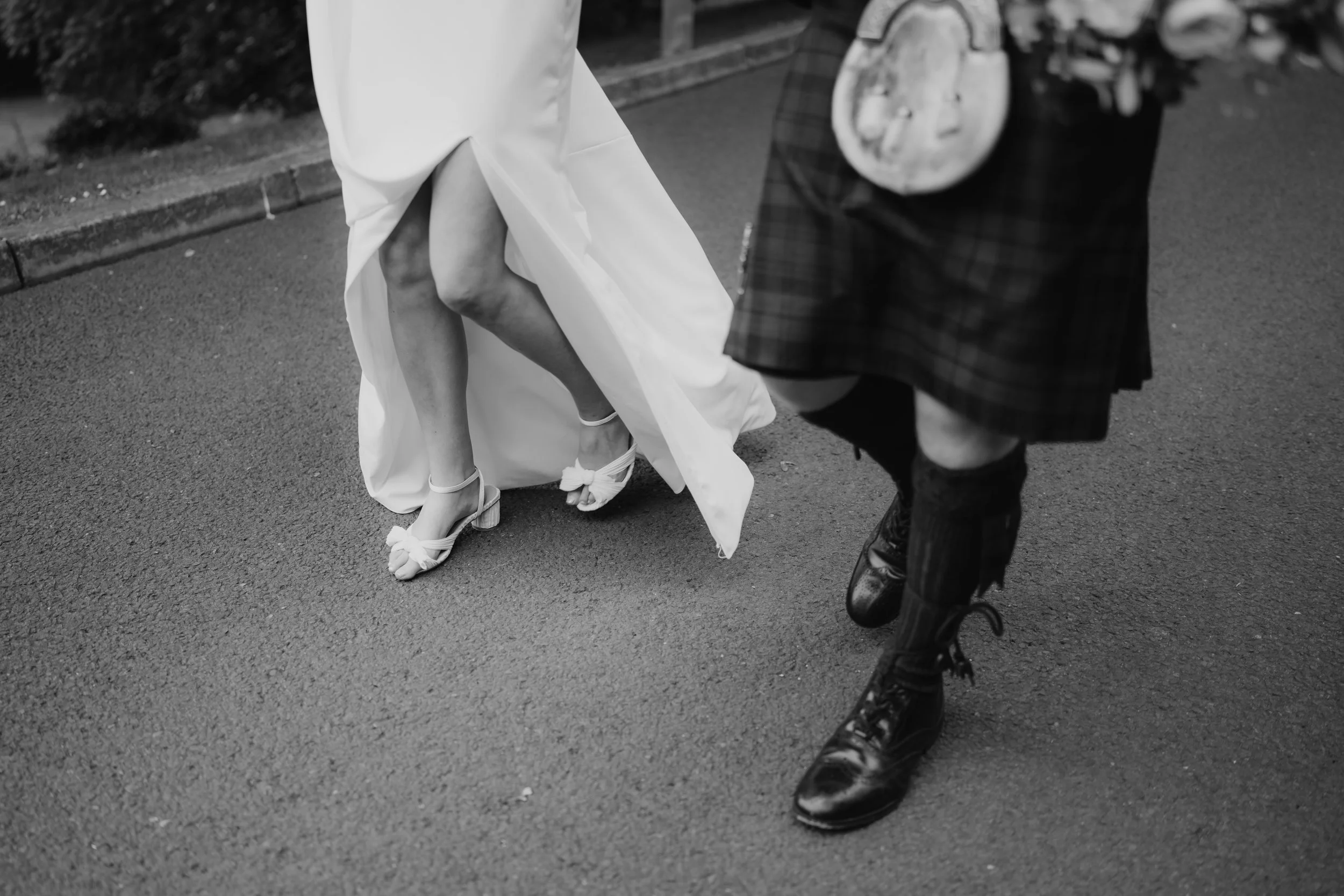 Close-up of a bride and groom's legs and feet during wedding, with the bride adjusting her shoe and the groom in traditional Scottish kilt, on a street