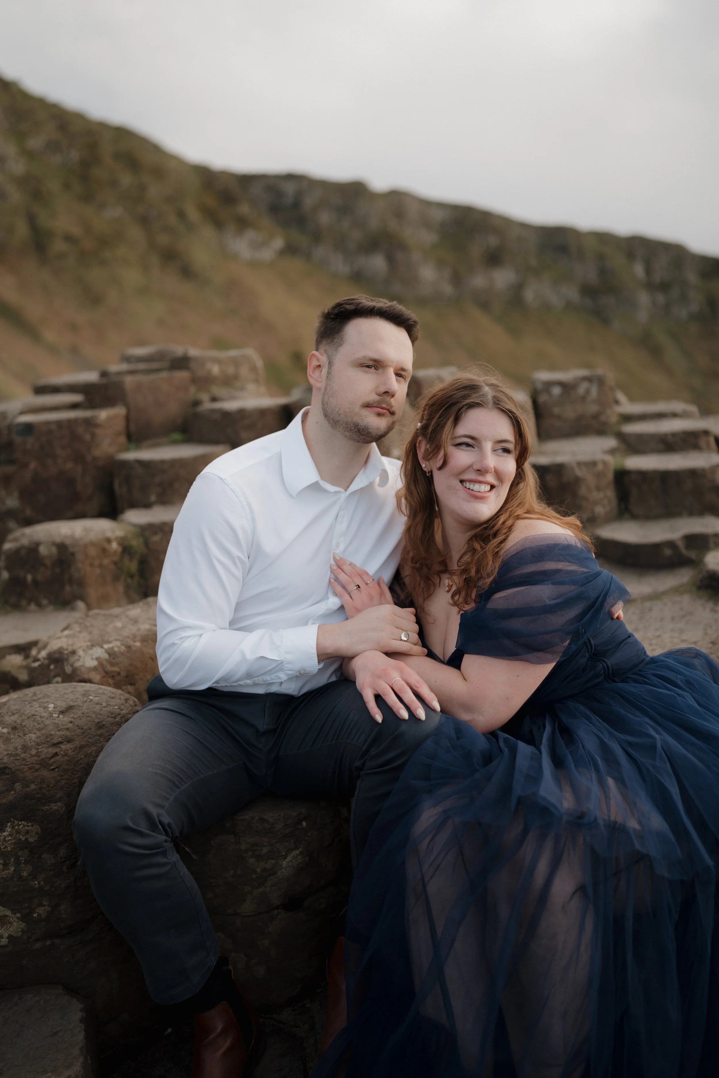 A couple sitting on rocks outdoors near a grassy hill, with the woman smiling and the man looking serious. The woman is wearing a navy blue dress and the man is dressed in a white shirt and dark pants.