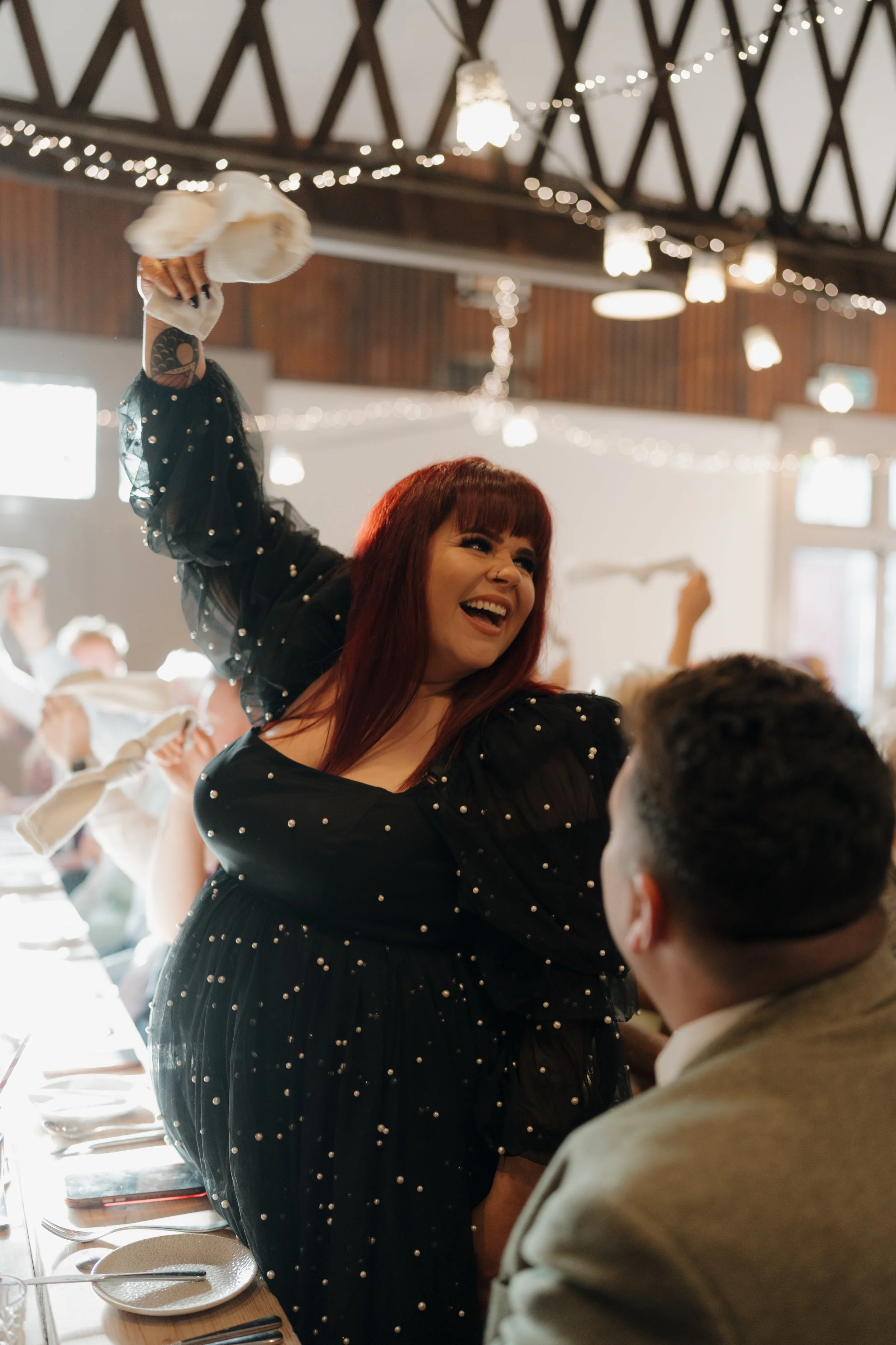 A woman with red hair and a black dress decorated with pearls is laughing and raising a napkin in celebration at a festive gathering in a decorated hall.