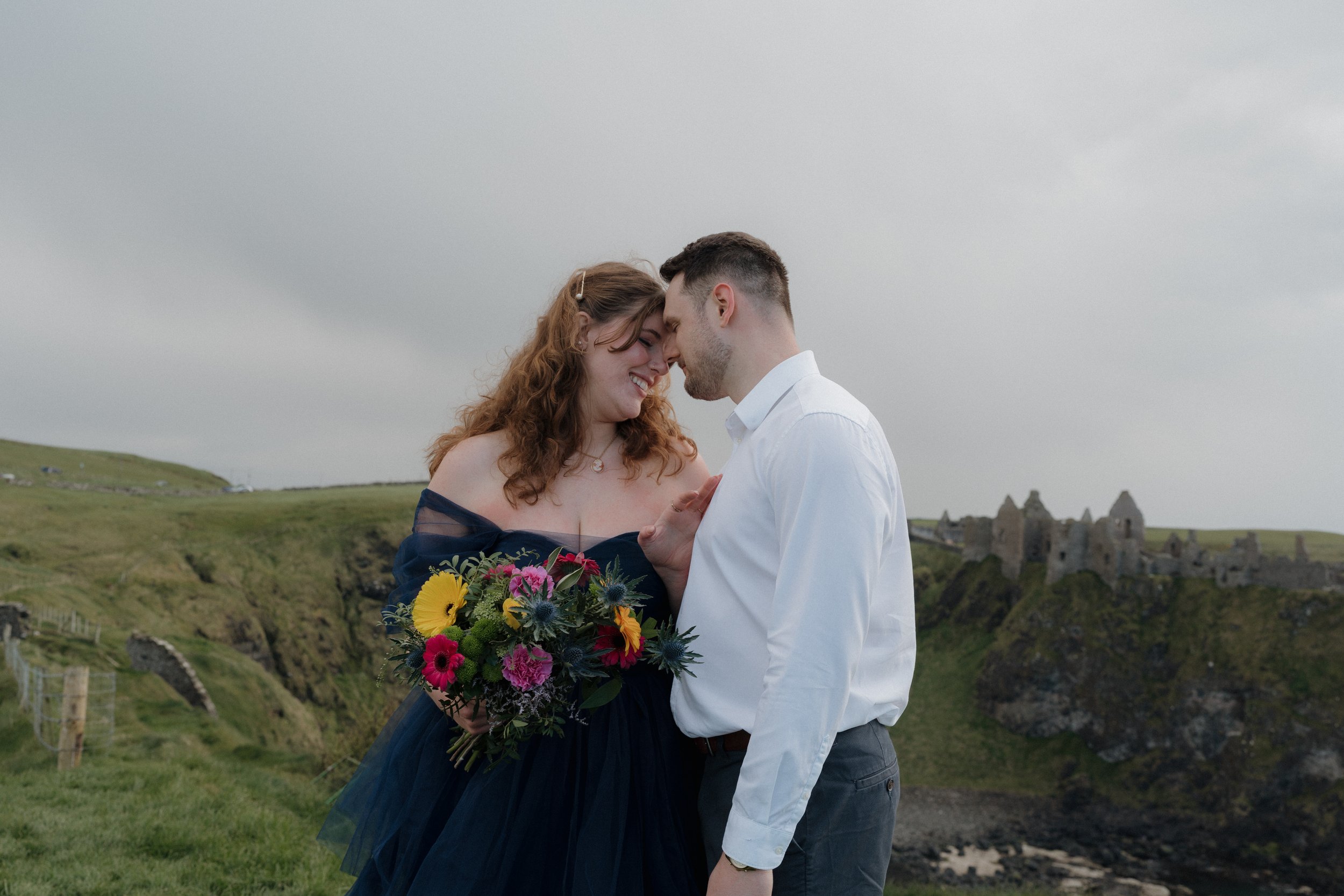 A smiling couple with their foreheads touching, standing outdoors on a cloudy day, with green hills and ruins of a castle in the background. The woman holds a colorful bouquet of flowers, and the man wears a white shirt.