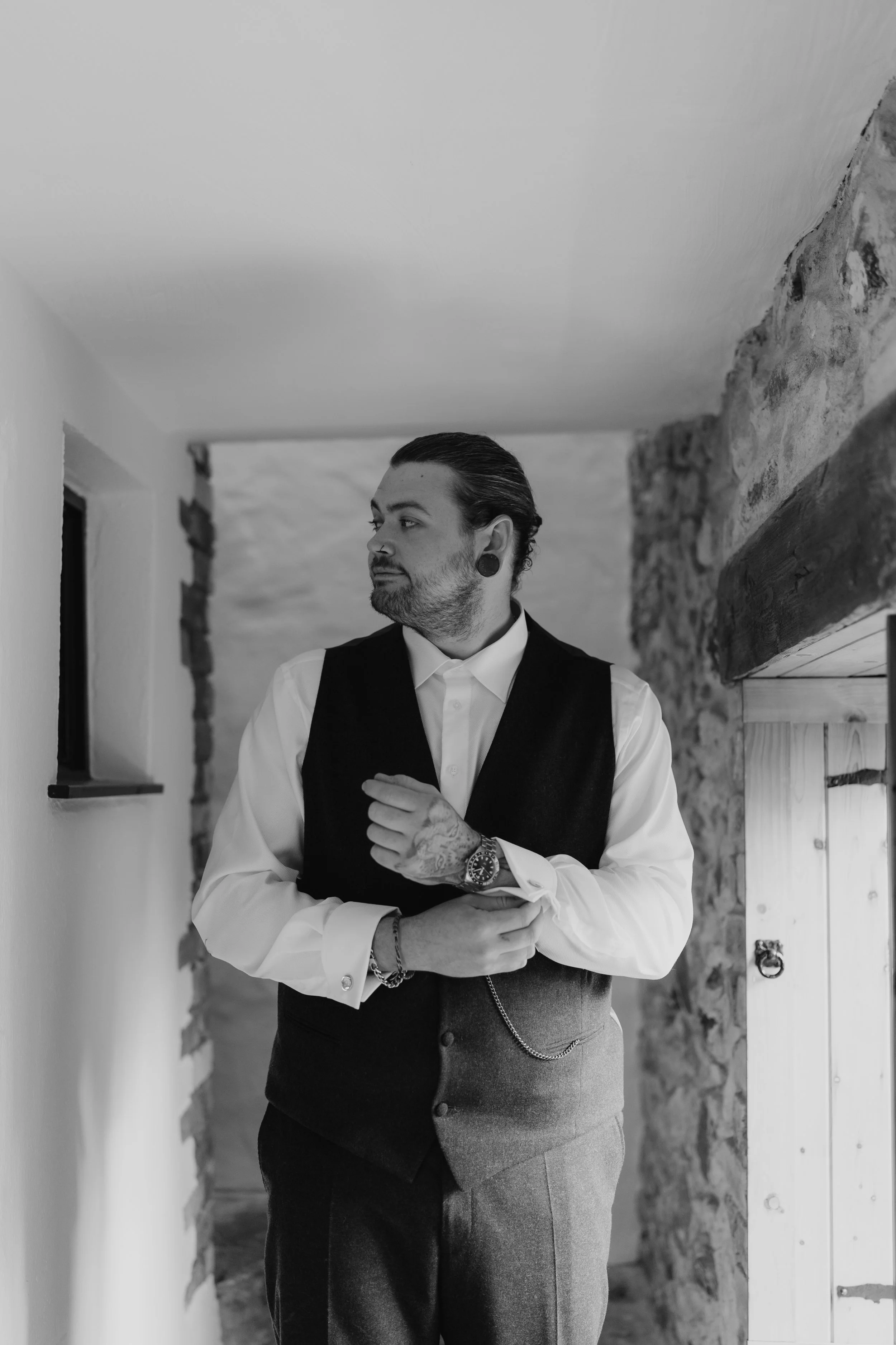 A man with slicked-back hair, tattoos, and earrings, wearing a white shirt, black vest, and gray pants, standing indoors in front of a stone wall and wooden beams.