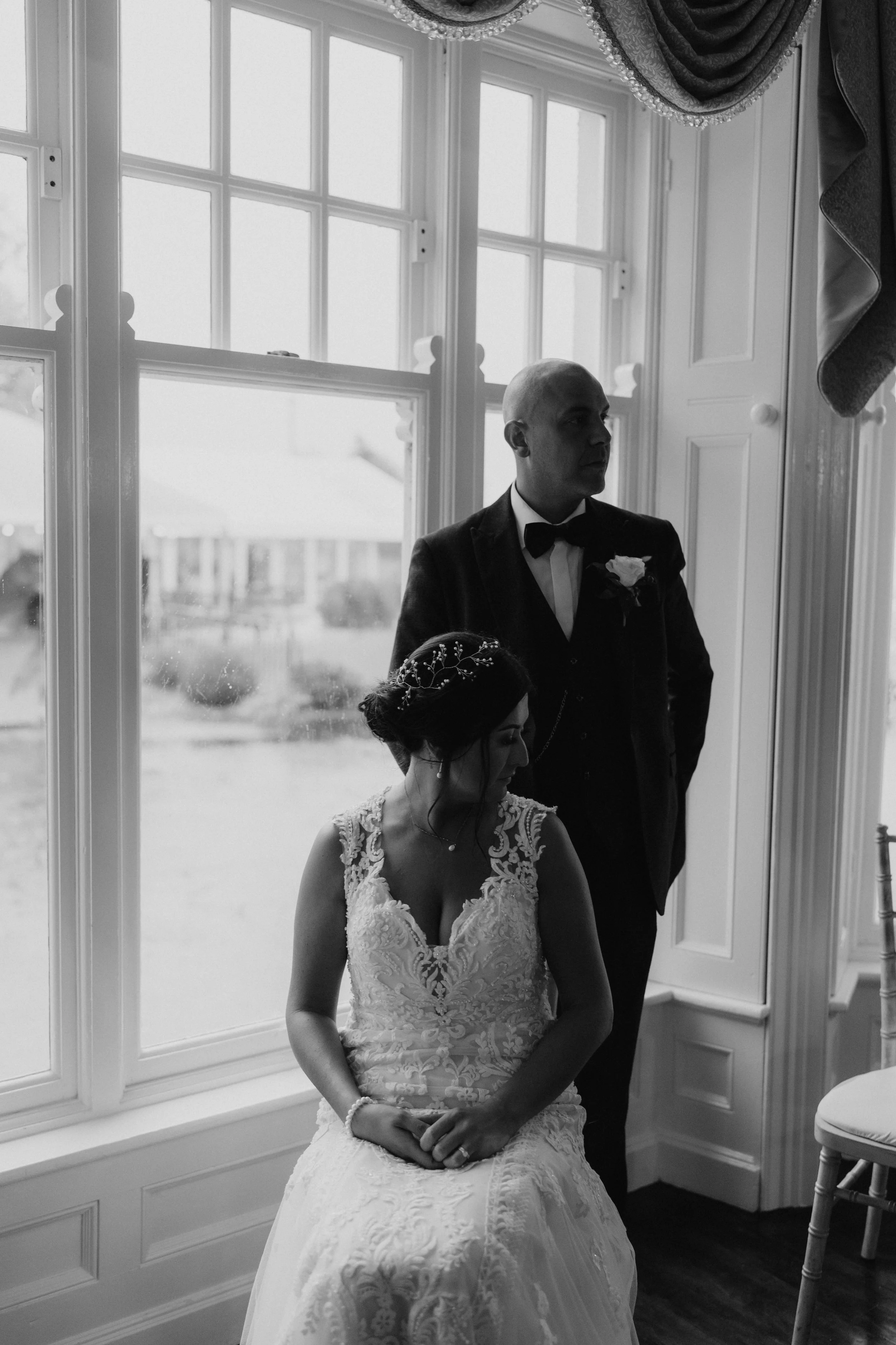 Black and white photo of a bride sitting on a window seat, wearing a lace wedding gown and a decorative headpiece, with a groom standing behind her near a large window with curtains.