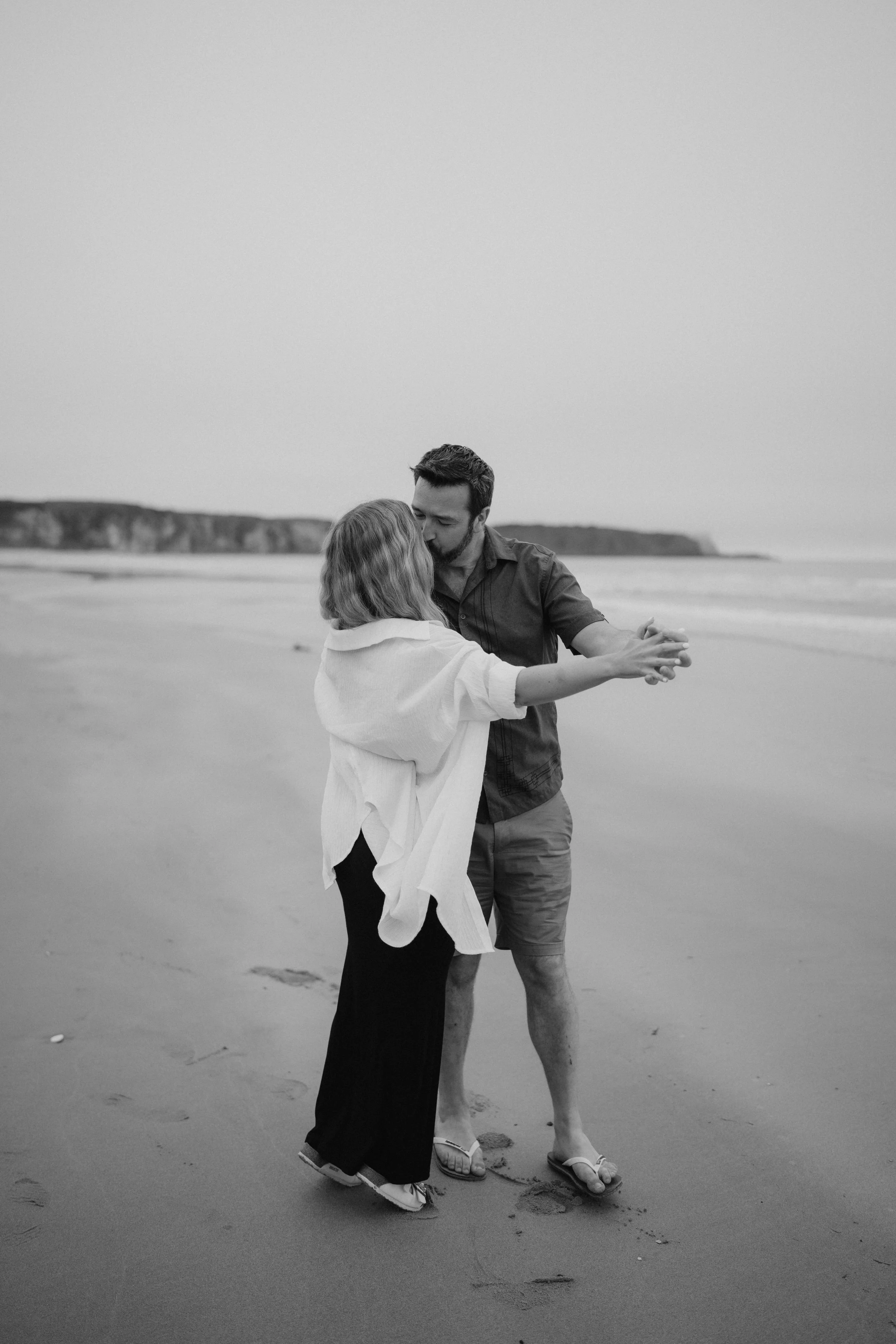 A couple dancing on a beach in black and white, with one person's hand resting on the other's shoulder and the man leaning in close to the woman.