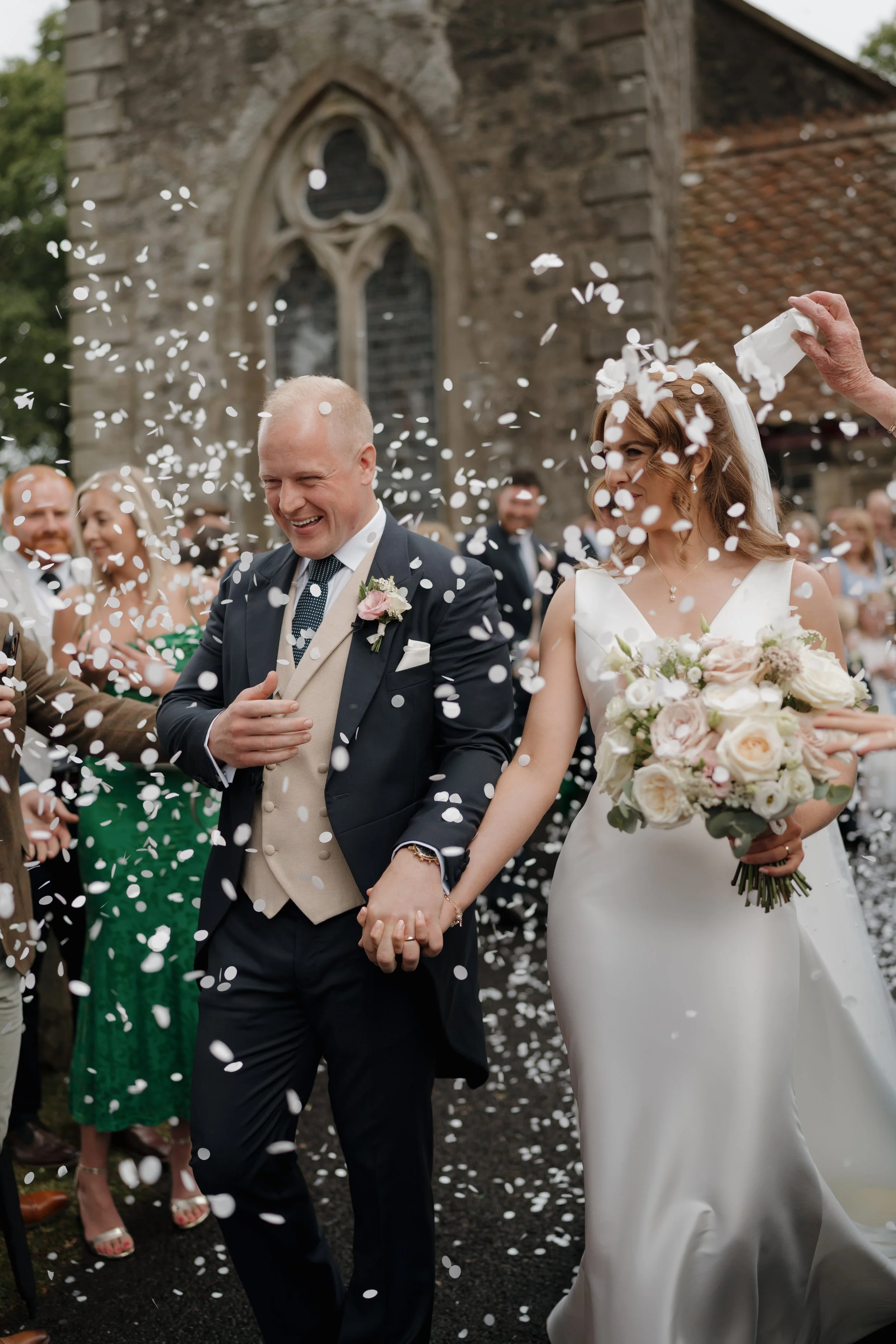 A newlywed couple is celebrating outside a church, surrounded by friends and family, as they walk hand in hand. The groom is in a dark suit with a beige vest, and the bride is in a white wedding dress holding a bouquet of flowers. White confetti is b