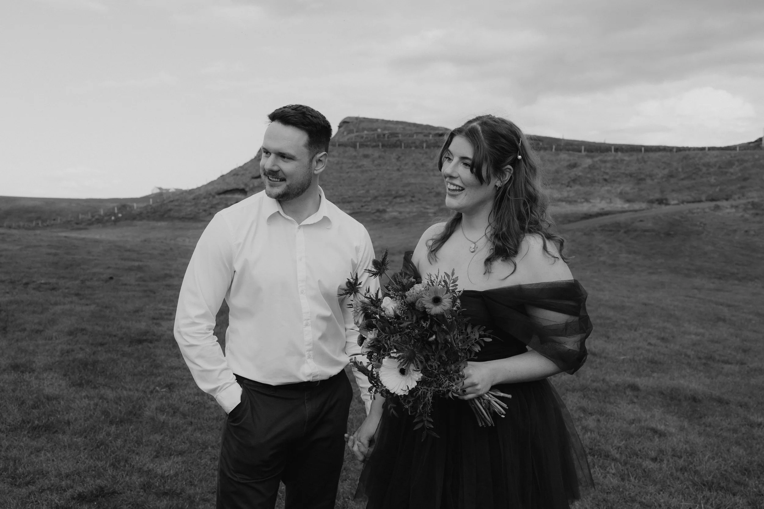 A black and white photograph of a smiling man and woman holding hands outdoors in a grassy field with a hill in the background. The woman is holding a bouquet of flowers and wearing a dark off-the-shoulder dress, while the man is dressed in a white s
