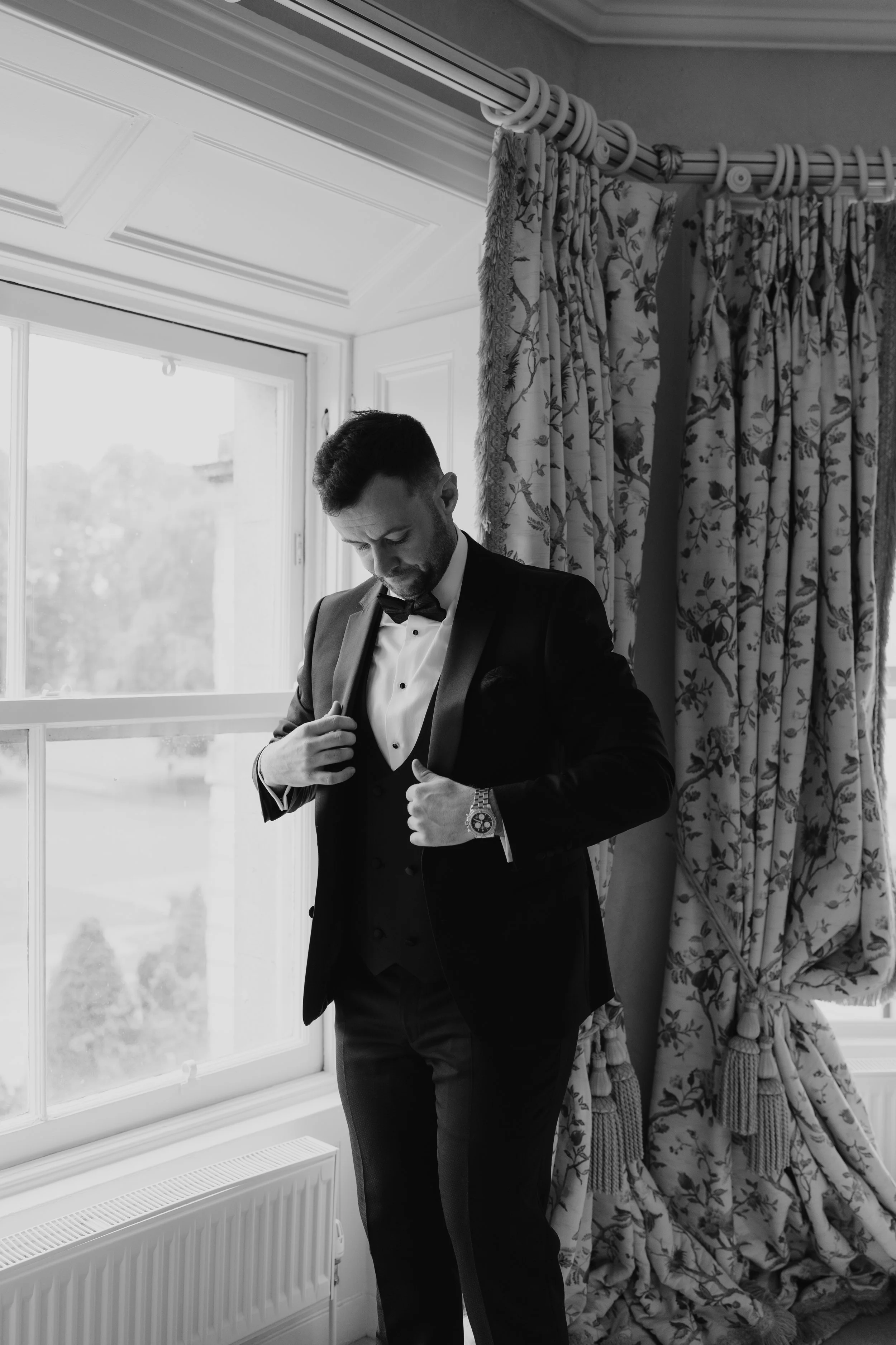 A man in a tuxedo adjusting his jacket near a window with patterned curtains, in black and white.