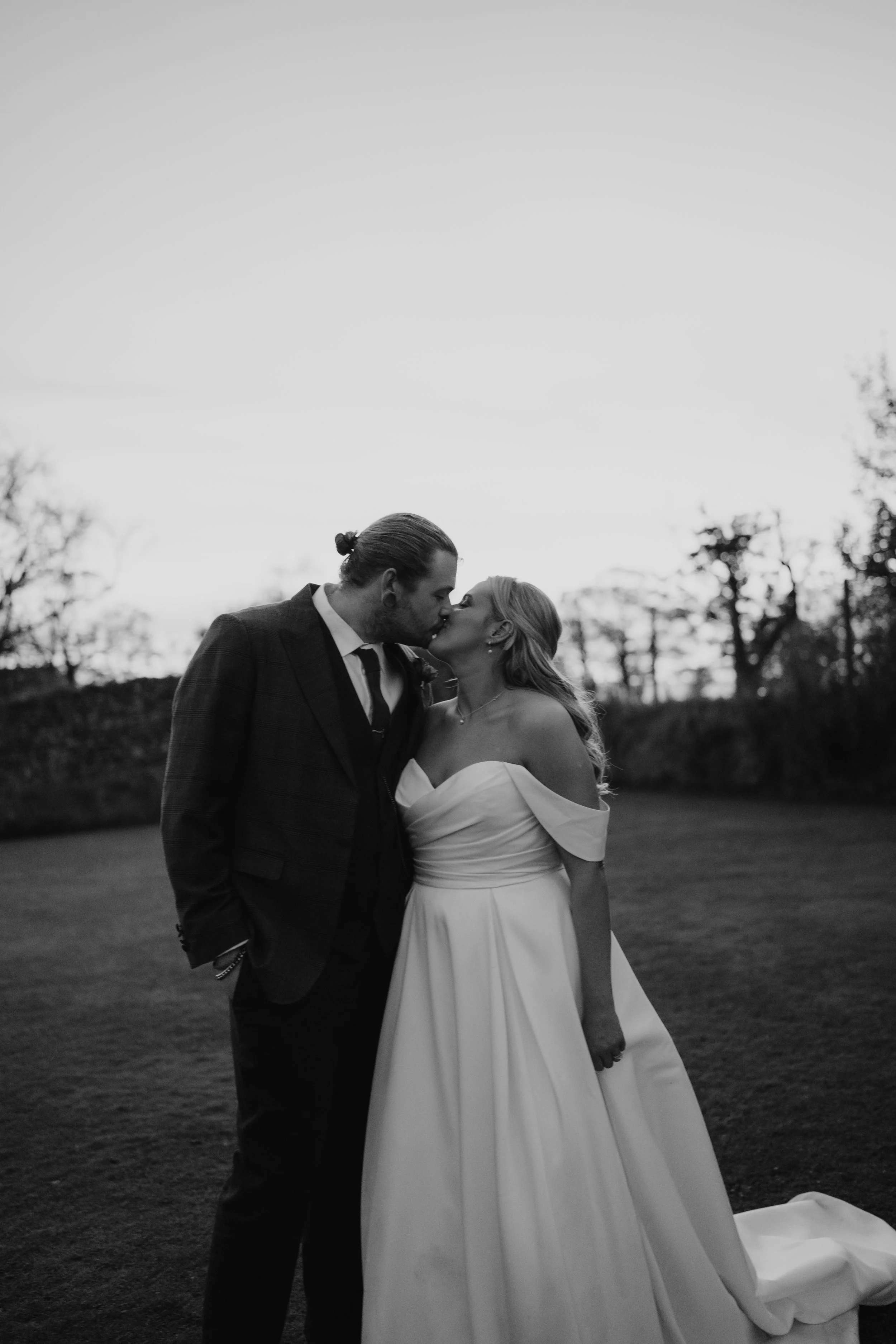 A black and white photo of a couple in wedding attire sharing a kiss outdoors at sunset.