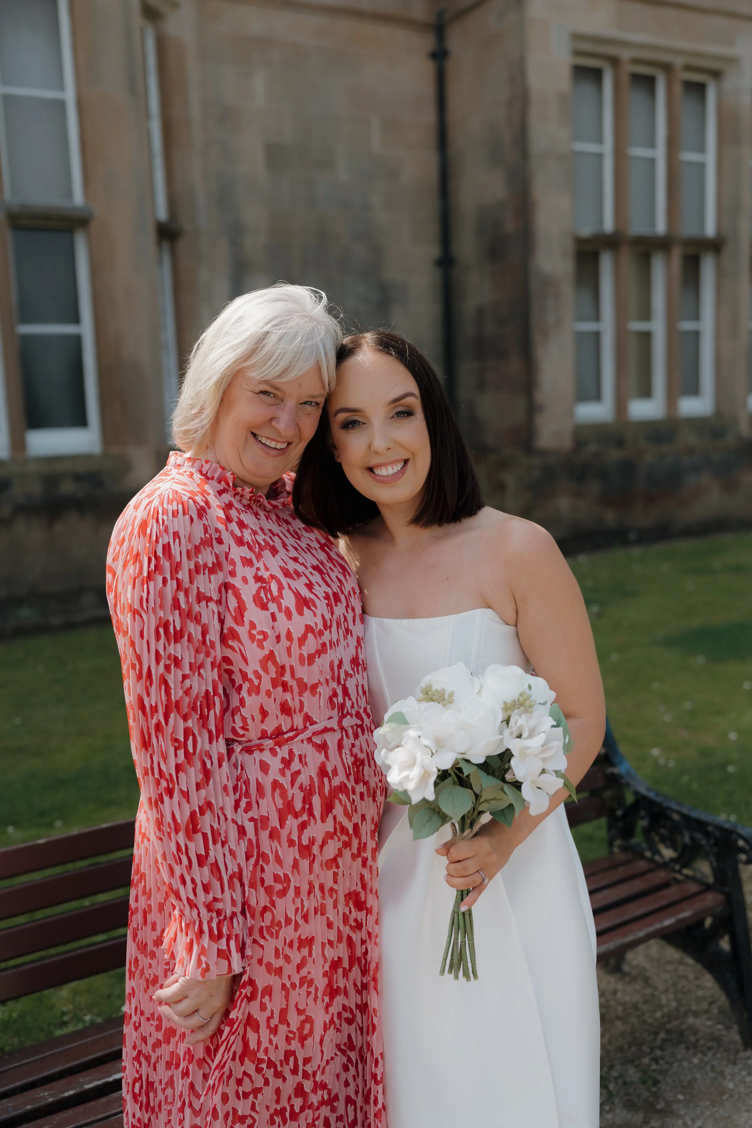 A woman in a white dress holding a bouquet of white flowers, standing next to an older woman in a pink and red patterned dress, both smiling happily outside in front of a stone building, Bangor Town Hall.