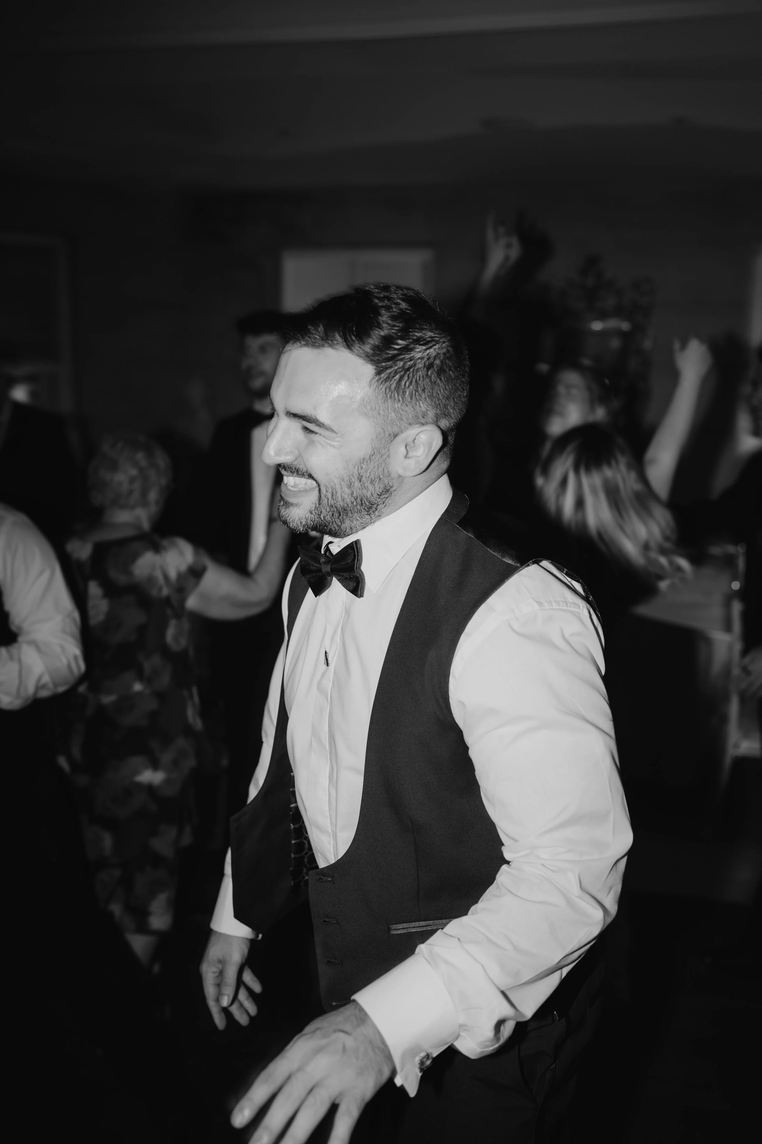 A man with a beard and short hair wearing a tuxedo and bow tie, smiling, dancing among others in a lively gathering.