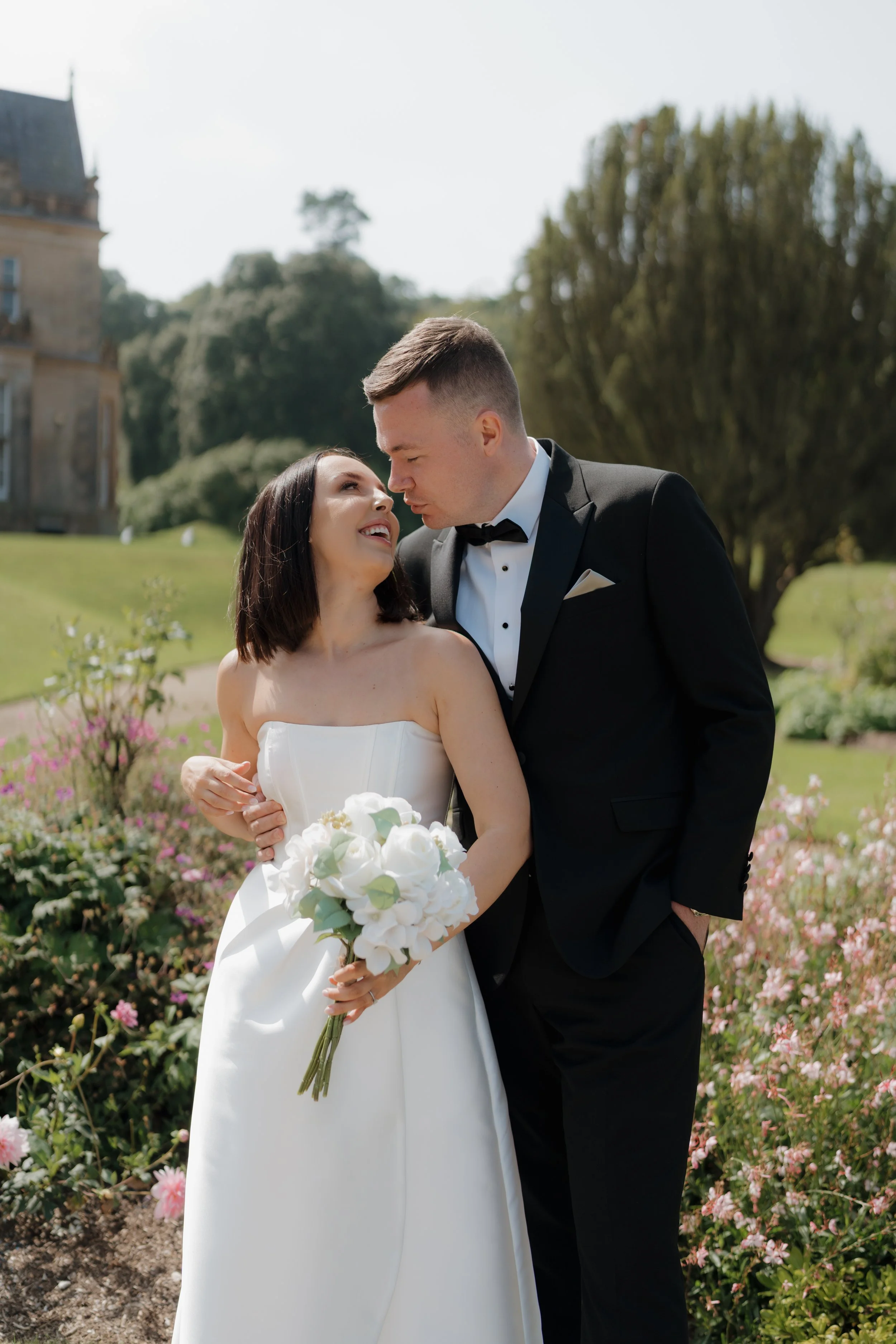A bride and groom in wedding attire embrace outdoors in a garden with flowers and a large estate in the background.