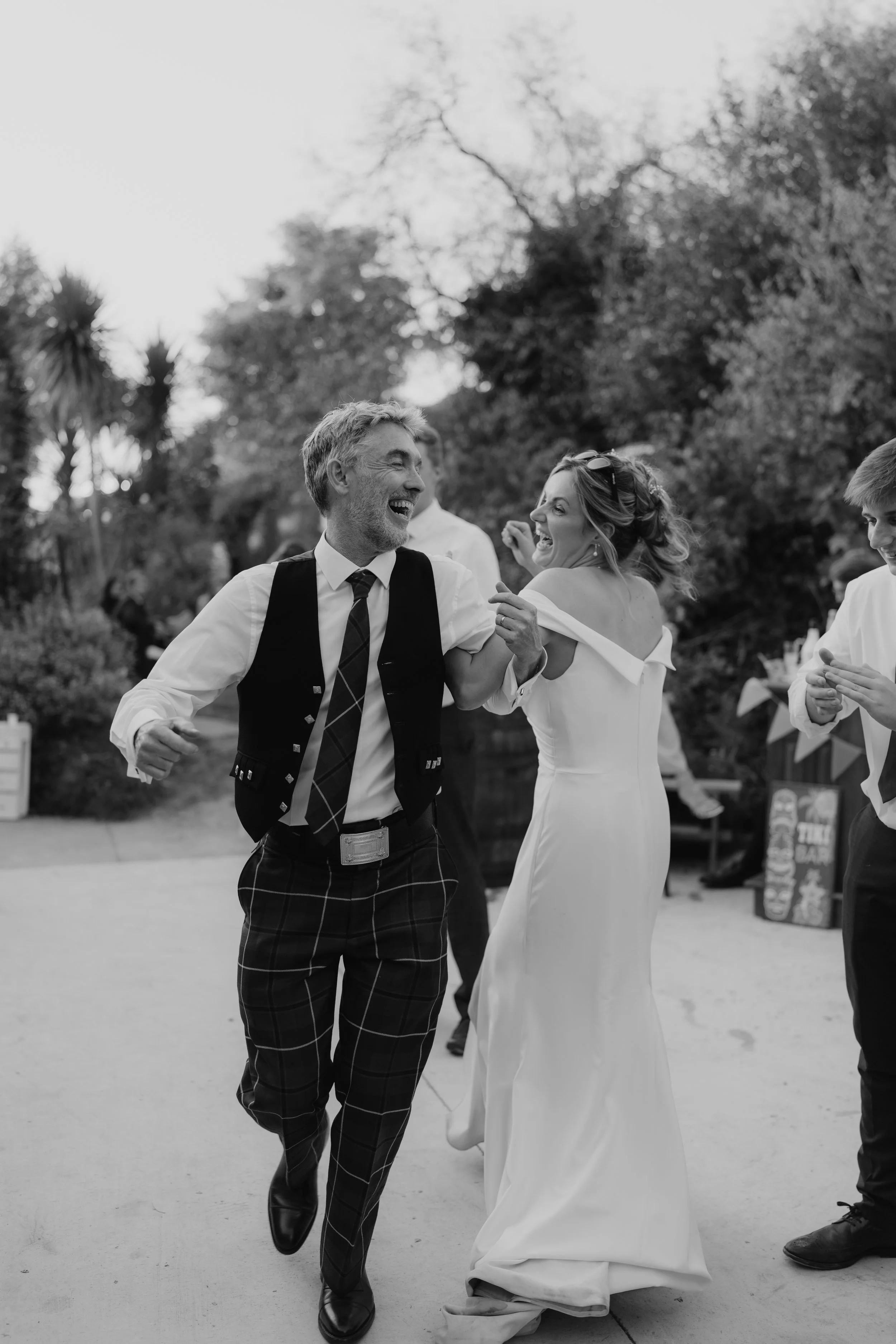 A black and white photo of a man and woman dancing outdoors. The man is wearing a vest, tie, and plaid pants, and the woman is in a white wedding dress. They are smiling and holding hands, enjoying the moment.