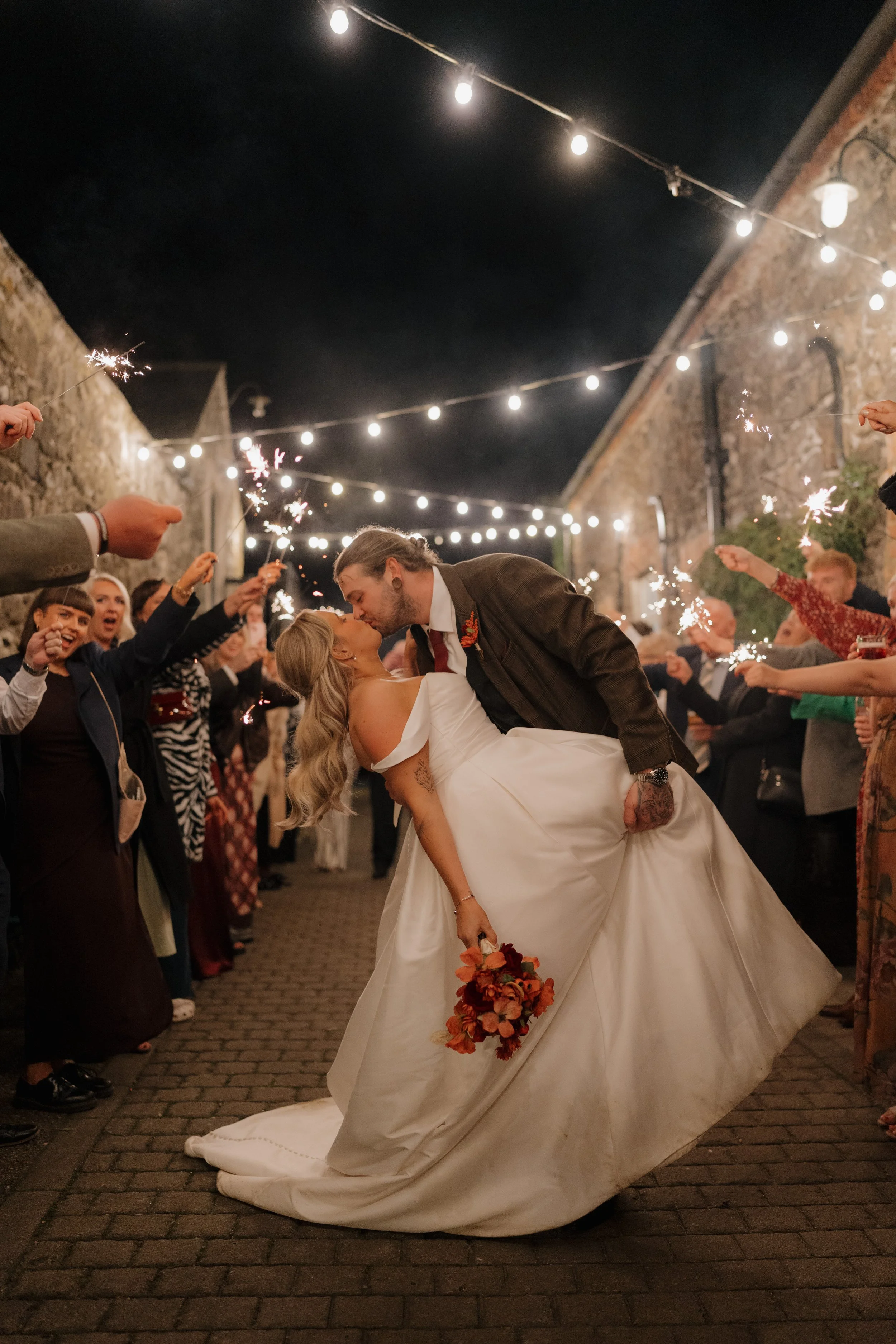 A newlywed couple shares a romantic moment during their wedding celebration, surrounded by guests holding sparklers under string lights in a cobblestone courtyard at night.